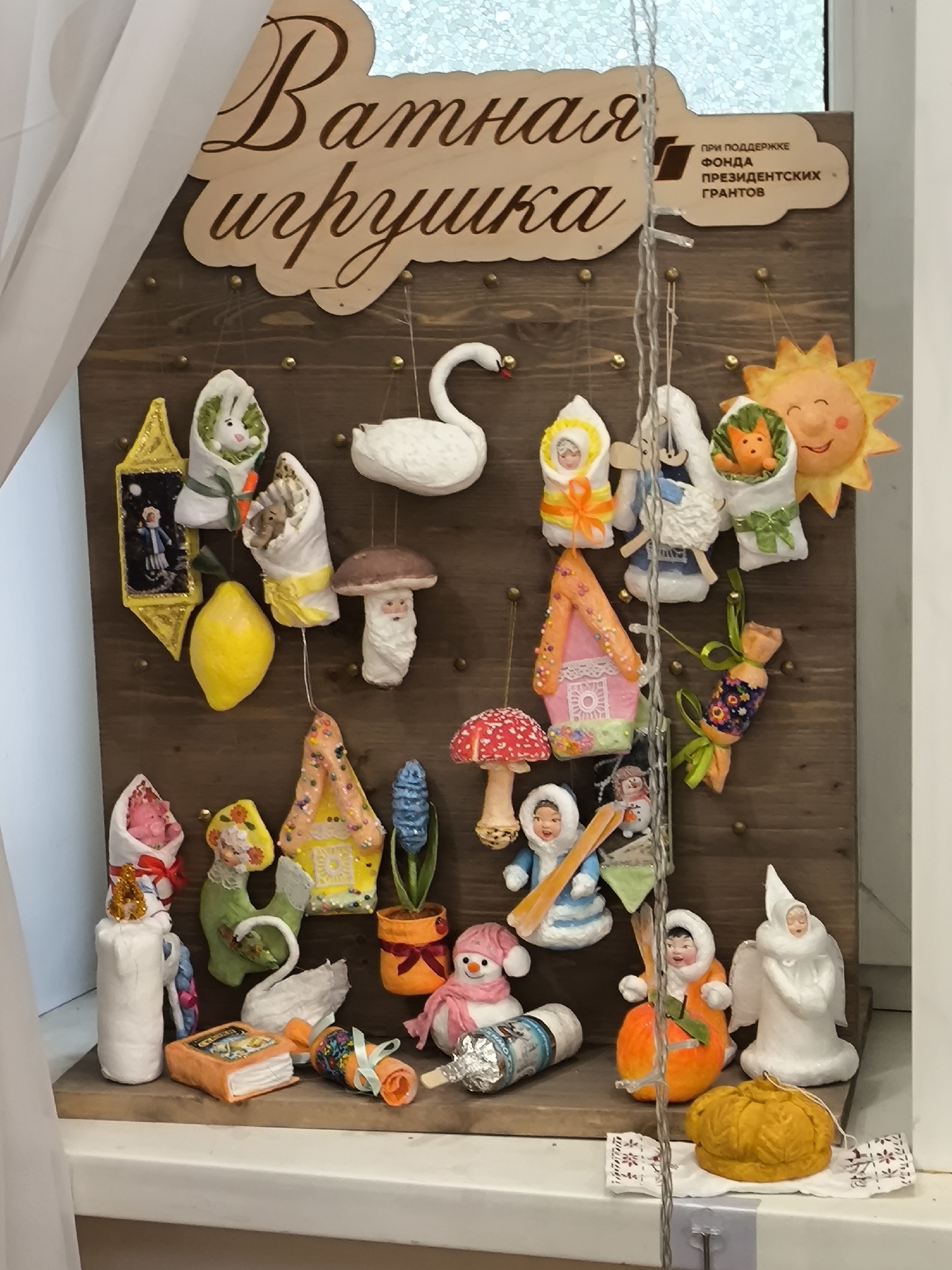 A display case contains a variety of handmade cotton toys on a wooden background. The toys are hung on pins on the wooden board, and some are placed on the shelf at the bottom. The toys include a swan, various figures wrapped in cloth, houses, mushrooms, and a sun. A sign above the display reads "Cotton Toy" in Russian, and another sign states "Supported by the Presidential Grants Fund". The display case is located in Moscow, Russia.