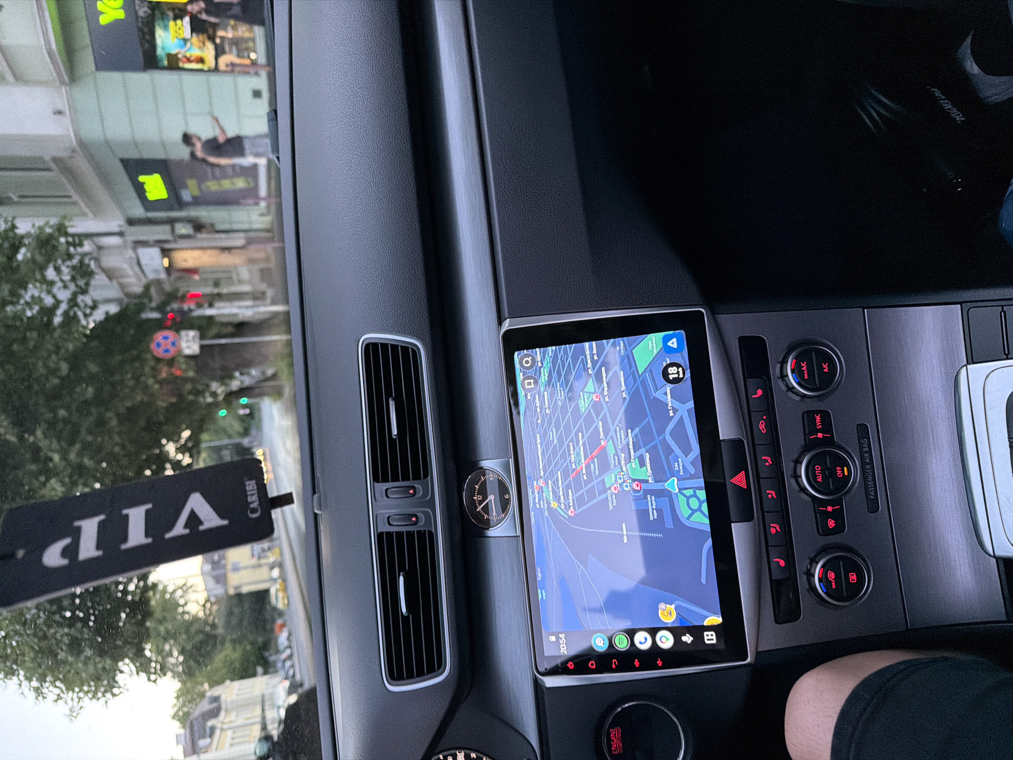 The image shows the interior of a car looking out at a street scene in Burgas, Bulgaria. Inside the car, a digital navigation screen displays a map of Burgas. The car's dashboard features climate controls and a clock. Outside the car, buildings line a street with traffic and pedestrians. A "VIP" air freshener hangs from the rearview mirror. The overall scene suggests a person driving or being driven through the city.