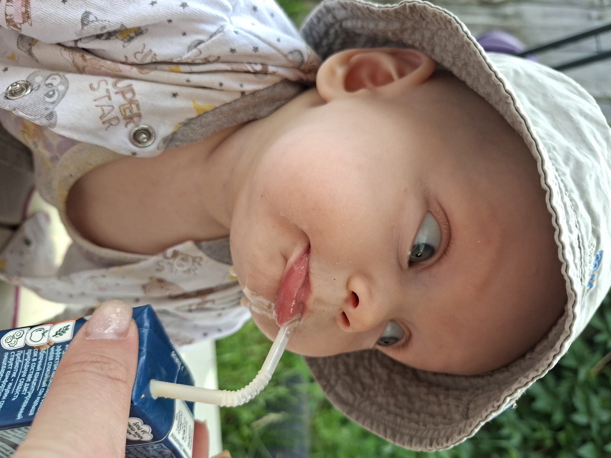 A close-up, eye-level shot shows a baby wearing a light-colored bucket hat and a white onesie with a grey trim, printed with cartoon animals and the words "SUPER STAR". The baby is being given a drink from a small blue juice box with a white straw. The baby's mouth is open, and juice is dribbling down its chin. A person's hand with a fingernail that has a glittery polish is holding the juice box. The background is out of focus and appears to be a green, leafy outdoor environment, possibly a garden or yard.