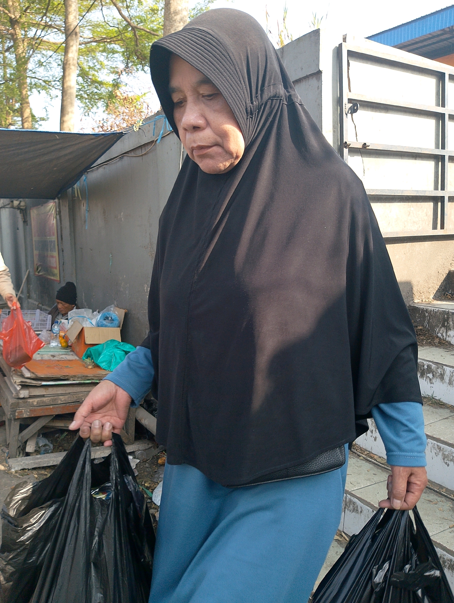 A medium shot captures an elderly Indonesian woman in Lembang, Indonesia, wearing a black hijab and a blue garment, carrying two black trash bags. The woman is positioned in the foreground, walking towards the viewer, her gaze directed downwards. Her right hand is extended, holding one of the trash bags by its handles. The background depicts a roadside stall with a tarp-covered roof, a man wearing a black beanie sitting at a table, and various items, including plastic bags and boxes. Beyond the stall, a concrete structure with metal railings and stairs is visible, suggesting an outdoor urban environment. The lighting suggests it is daytime, with sunlight casting shadows on the ground and the woman's clothing.