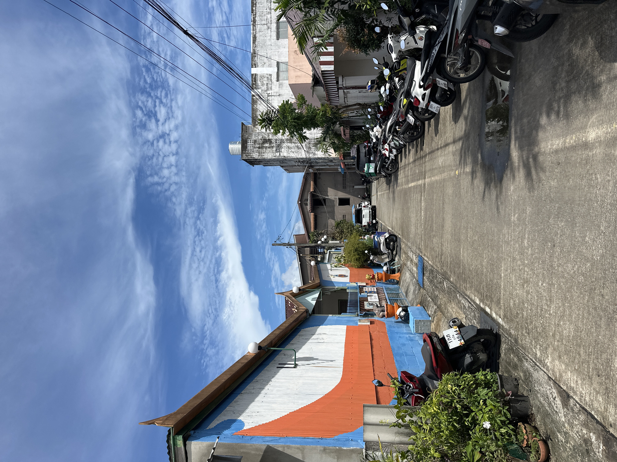 A street scene is captured in an outdoor, daylight setting. The perspective looks down a paved road lined with buildings on either side and parked motorcycles. The building on the left is painted with a colorful design of blue, white, and orange wavy lines. The building on the right has a more muted facade, with some appearing to be shops or residences. Several motorcycles are parked along the right side of the street. The sky above is blue with scattered white clouds. There is a visible utility pole with wires running across the upper portion of the image. The overall impression is of a residential or commercial street in a tropical or subtropical climate.
