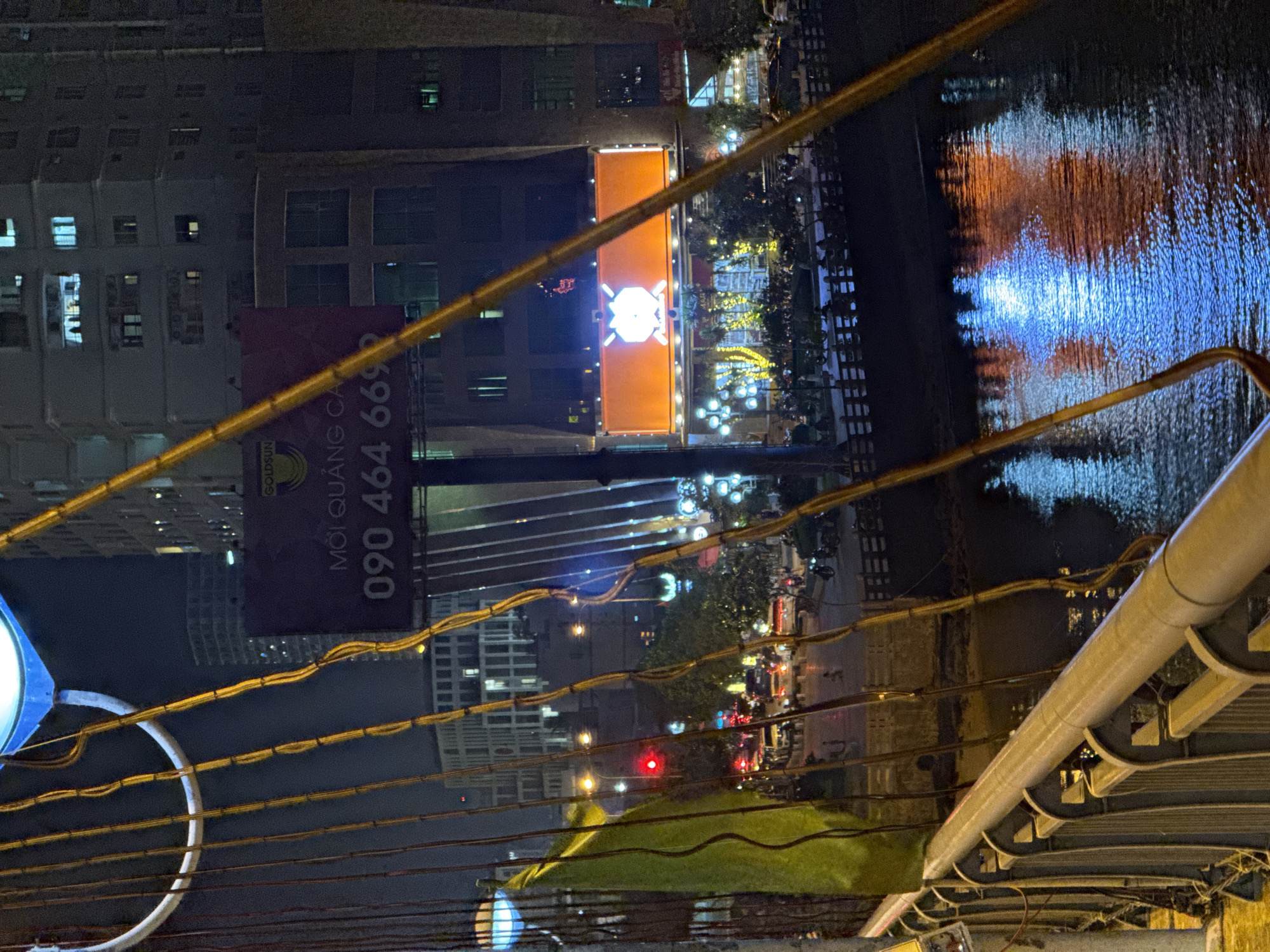 The image captures a nighttime view of the central area of Nha Trang, Vietnam, likely from a bridge overlooking a body of water. In the foreground, a railing of a bridge is visible, with cables and wires strung along its side. A green flag is draped over a railing.
Dominating the mid-ground is a large billboard for "GOLDSUN" displaying "MỞ QUÀNG CA 090 464 6692". Behind the billboard, a building with illuminated windows is seen. Further back, another large, brightly lit orange advertisement with a white geometric symbol is displayed on a building facade.
The scene is set at night, with the ambient light from buildings, streetlights, and the advertisements casting reflections on the water below. A street with traffic, including cars and motorcycles, is visible, with a red traffic light illuminating the scene. People can be seen walking on the sidewalk or near the water's edge. The overall impression is a vibrant urban nighttime environment.
