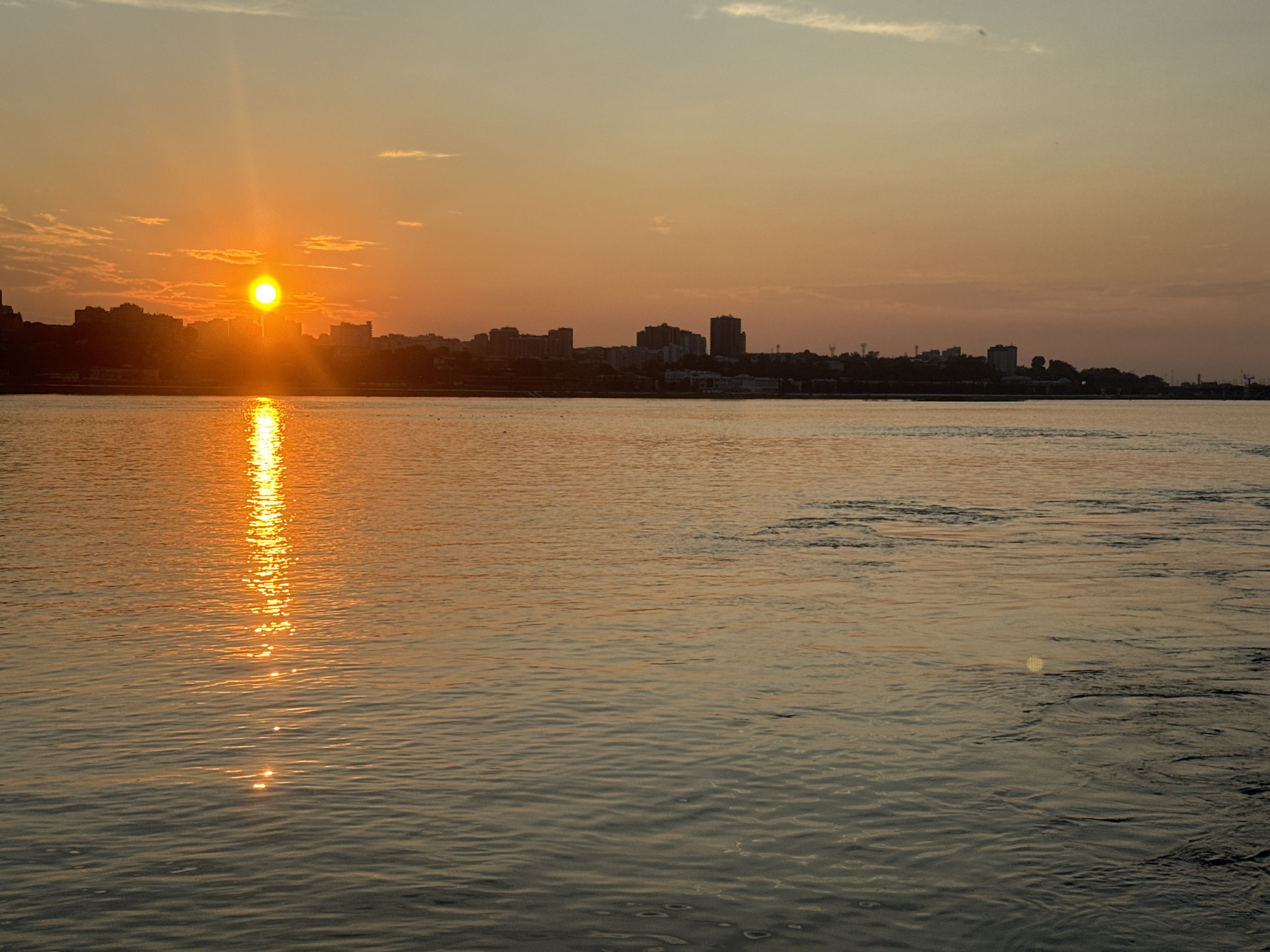 The image depicts a sunset over a body of water, likely a river, with a city skyline in the background. The sun, a bright orange orb, is low in the sky on the left side of the frame, casting a strong reflection on the water. The sky is a gradient of orange and yellow, with some wispy clouds. The city skyline consists of buildings of various heights, silhouetted against the colorful sky. The water is rippled, showing the effect of light on its surface. The scene appears to be a calm and serene evening. Considering Irkutsk, Russia, this view is likely of the Angara River, which flows through the city. The presence of the city skyline suggests a viewpoint from across the river, looking towards the urban center of Irkutsk. There are no people or specific actions depicted.
