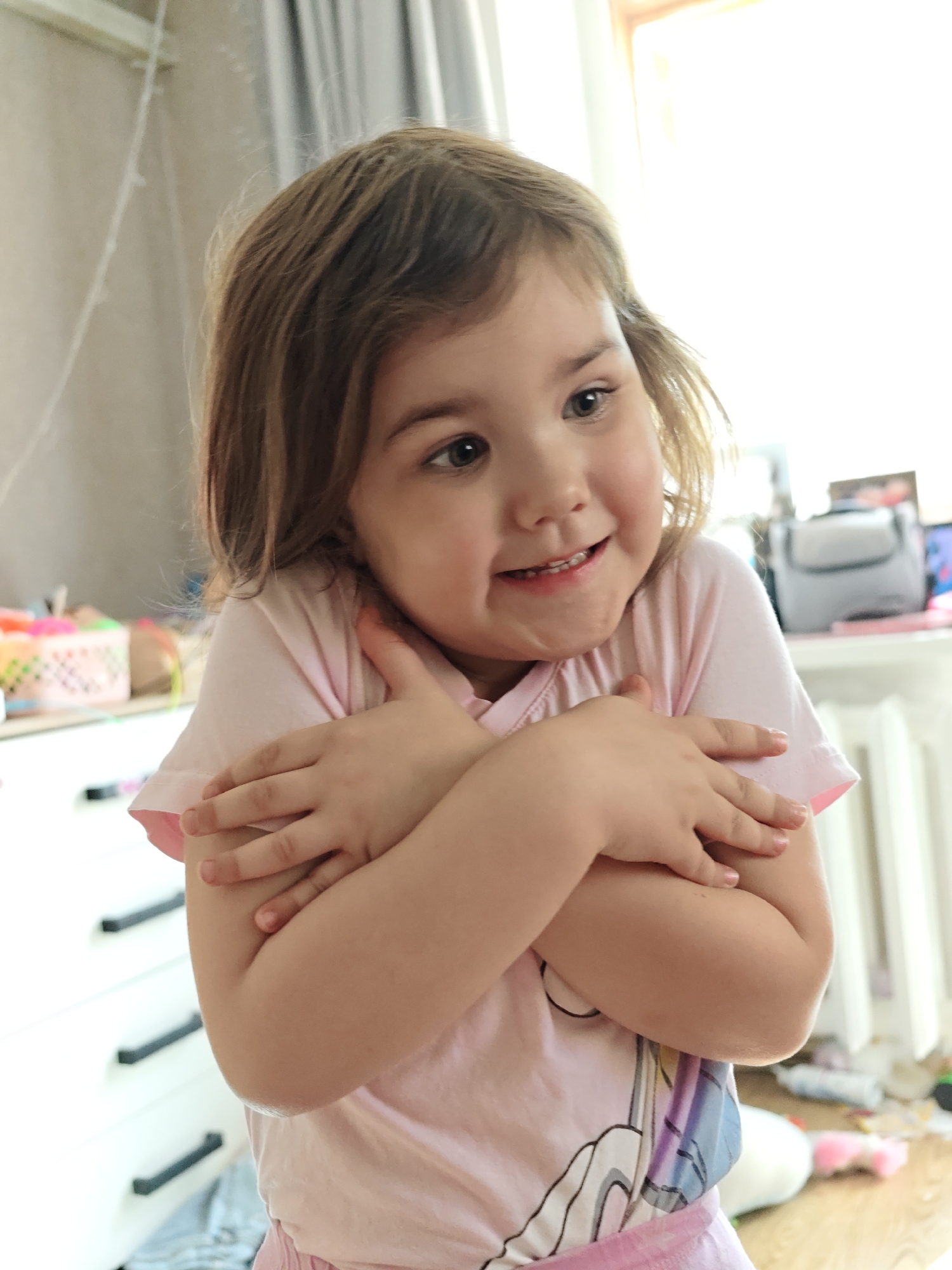 A young child with brown hair and light skin stands indoors, wearing a light pink t-shirt with a cartoon unicorn design and light pink pants. The child is smiling slightly and has both arms crossed tightly across their chest. In the background is a white dresser with black handles, and a window letting in bright light. Behind the child and dresser is a desk/table with various objects on it, including a grey case, a pink basket, and other items. The location appears to be a child's bedroom, possibly in a residence in Chaikovsky, Russia.