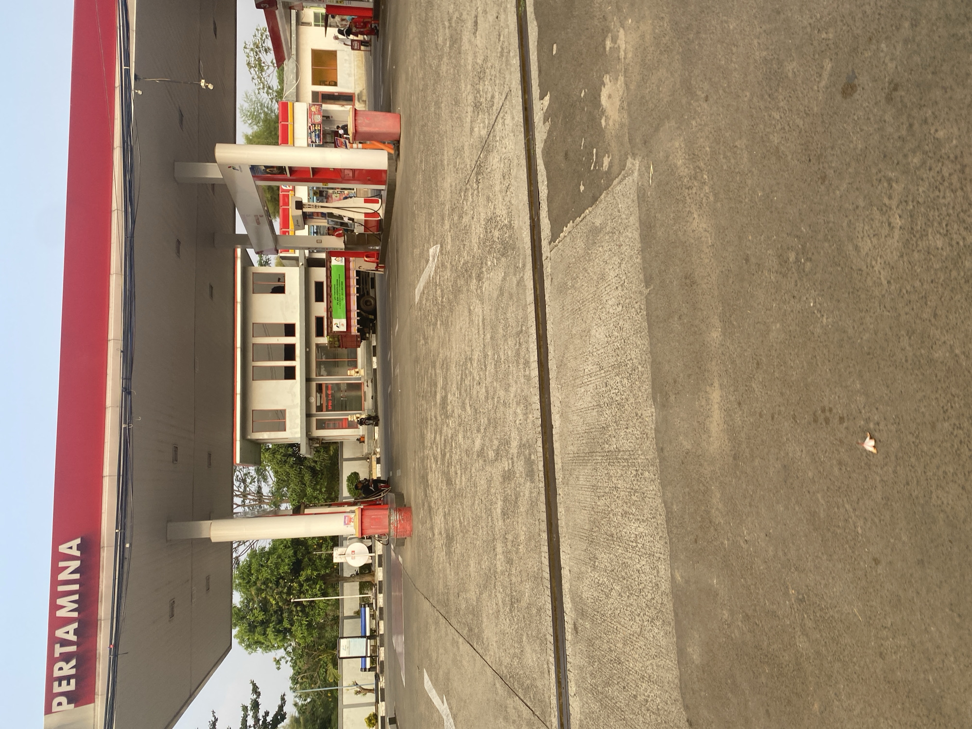 The image shows a Pertamina gas station in Depok, Indonesia. The gas station has a large red and white canopy over multiple fuel pumps. A white building with multiple windows is visible behind the pumps. To the left of the building, there is a truck with a green banner on its side. A few people are present at the gas station, including a person near the fuel pumps and a couple of individuals near the building. The ground is paved with concrete. The sky is clear and a few trees are visible in the background.