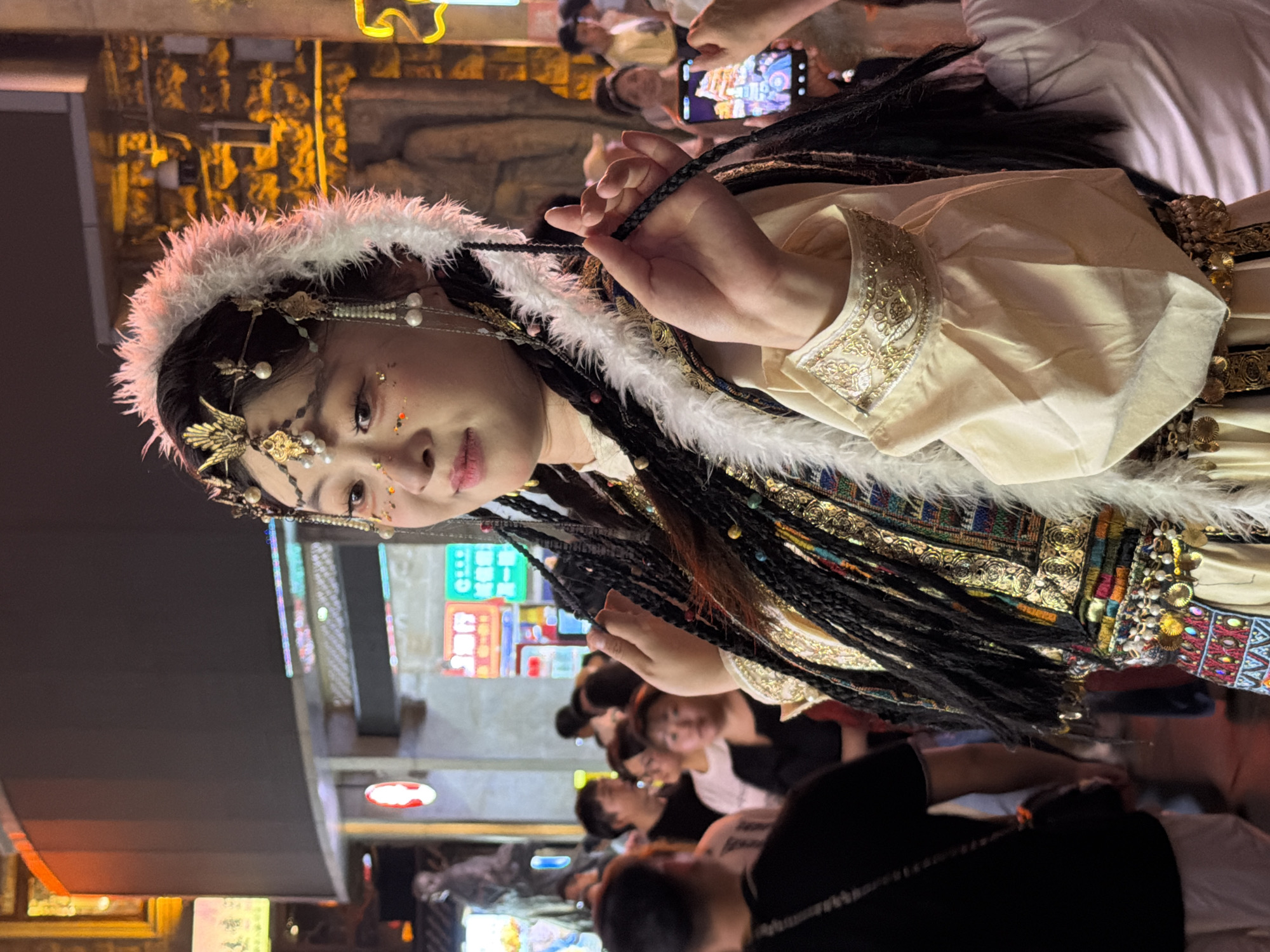 The image shows a woman dressed in traditional clothing, possibly a costume, with a fur-lined headdress and elaborate jewelry adorning her face and hair. She has long, braided hair with colorful beads woven in. Her outfit is ornate, featuring gold embroidery and patterned fabric. She is holding a braided cord in her right hand, which is part of her costume.
The scene appears to be a bustling night market or street event, given the presence of neon signs in the background. Several other people are visible, some looking at the woman, and one person in the upper right is holding a smartphone, possibly taking a photo or video of her. The lighting suggests it is nighttime. The overall atmosphere seems festive and lively. The background shows illuminated signs with Chinese characters and a building structure.
The woman's expression is engaging, and the overall composition focuses on her and her detailed attire. The setting hints at a cultural performance or a special event happening in a public space in Yuzhong District, China, at night.