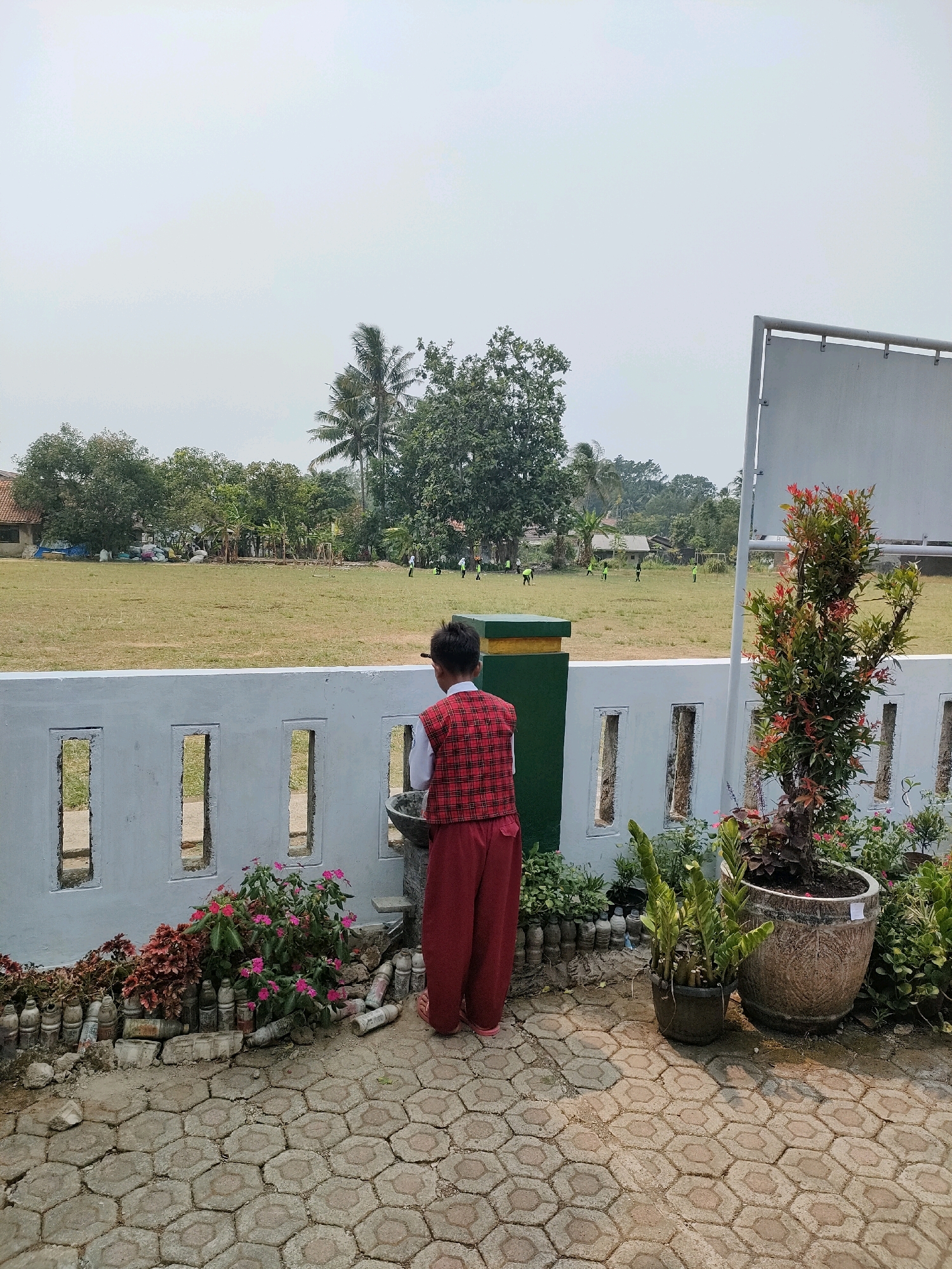 A young boy, wearing a red plaid vest over a white shirt and red pants, stands with his back to the camera, facing a drinking fountain in what appears to be an outdoor schoolyard in Bojong, Indonesia. He is positioned in the foreground, to the left of the frame. Behind him, a white, low-lying fence with rectangular cutouts separates the foreground from a large, grassy field where several other children in light-colored shirts and dark pants are scattered, seemingly engaged in some sort of activity. Trees, including palm trees, and some buildings are visible in the background, suggesting a rural or suburban setting. Potted plants, including a tall plant with red-tipped leaves and a smaller potted plant with green foliage, are situated to the right of the boy, near the fence. The ground in the foreground is paved with hexagonal interlocking concrete blocks. The sky is overcast.