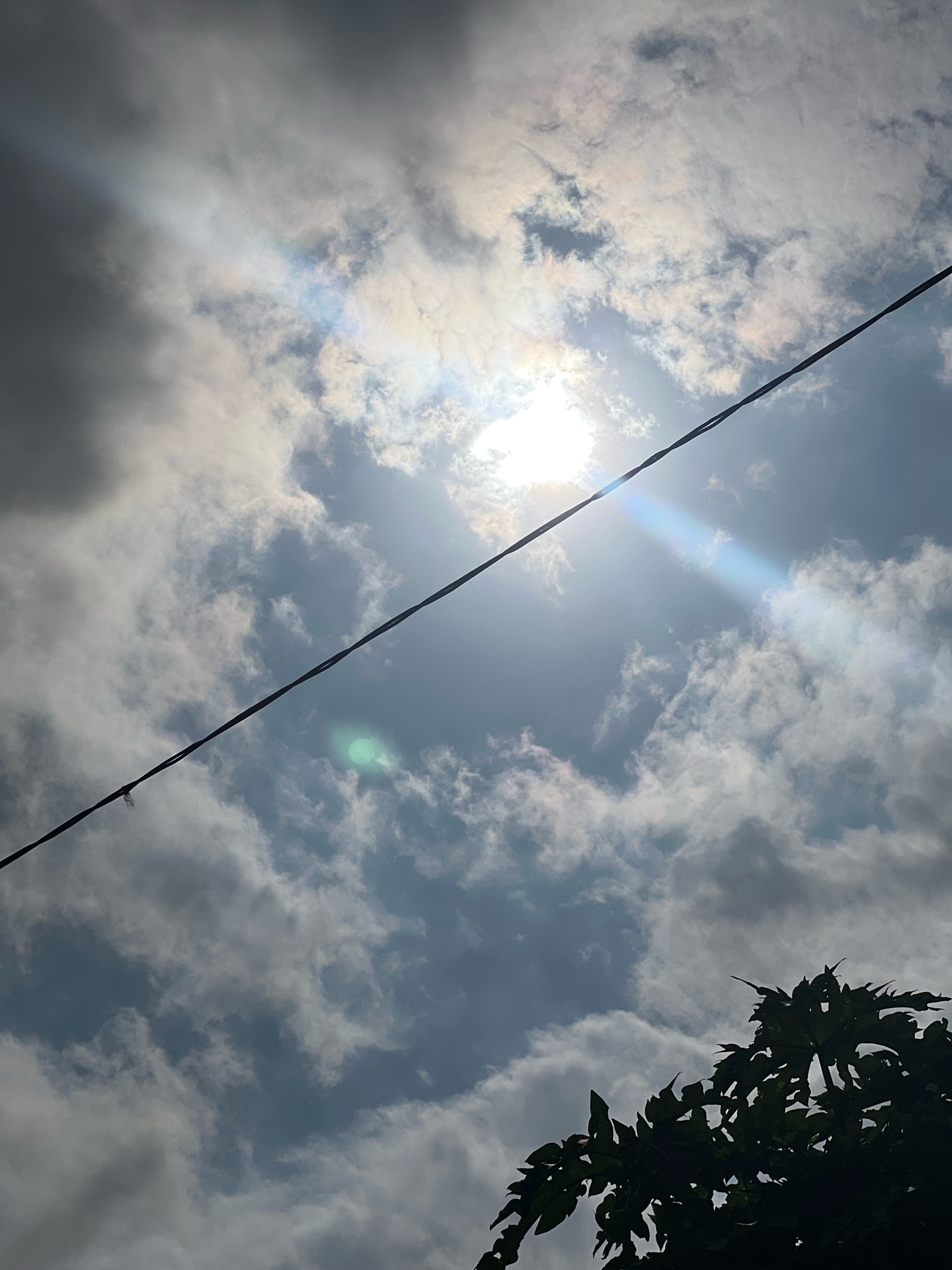 The image shows a view looking up at a cloudy sky in Denpasar, Indonesia. A dark, thin wire stretches diagonally across the frame from the bottom left to the top right. The sun is visible through breaks in the clouds, creating bright flares and a faint rainbow effect in the sky. In the bottom right corner, the silhouette of a leafy tree is visible against the sky. The clouds are varied in density, with some appearing dark and heavy and others thin and wispy. The overall impression is of a slightly overcast day with intermittent sunshine.