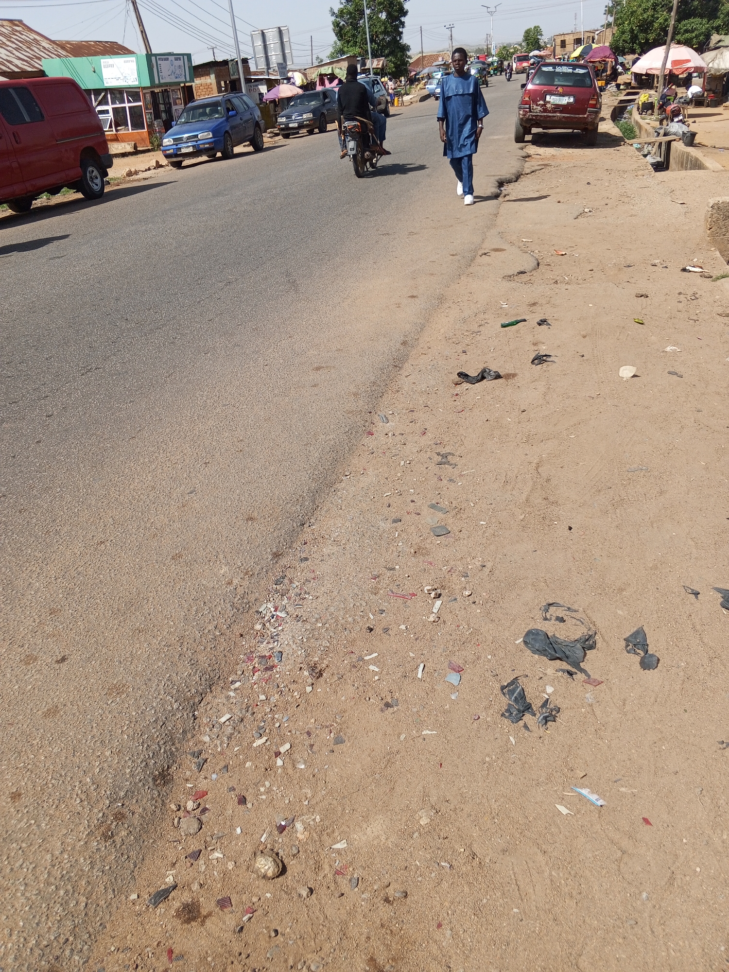 The image depicts a street scene in Bukuru, Nigeria. A paved road is visible, with a sandy shoulder on the right side littered with debris, including plastic bags and fragments of various materials. Several vehicles are parked along the roadside, including a red van on the left, a blue car, and a red car further down the road. A man in a blue outfit is walking on the road toward the viewer. Another person is riding a motorbike ahead of him. In the background, there are buildings, trees, and people, suggesting a populated area. Some market stalls with parasols can be seen in the distance. The sky is clear, indicating a sunny day.