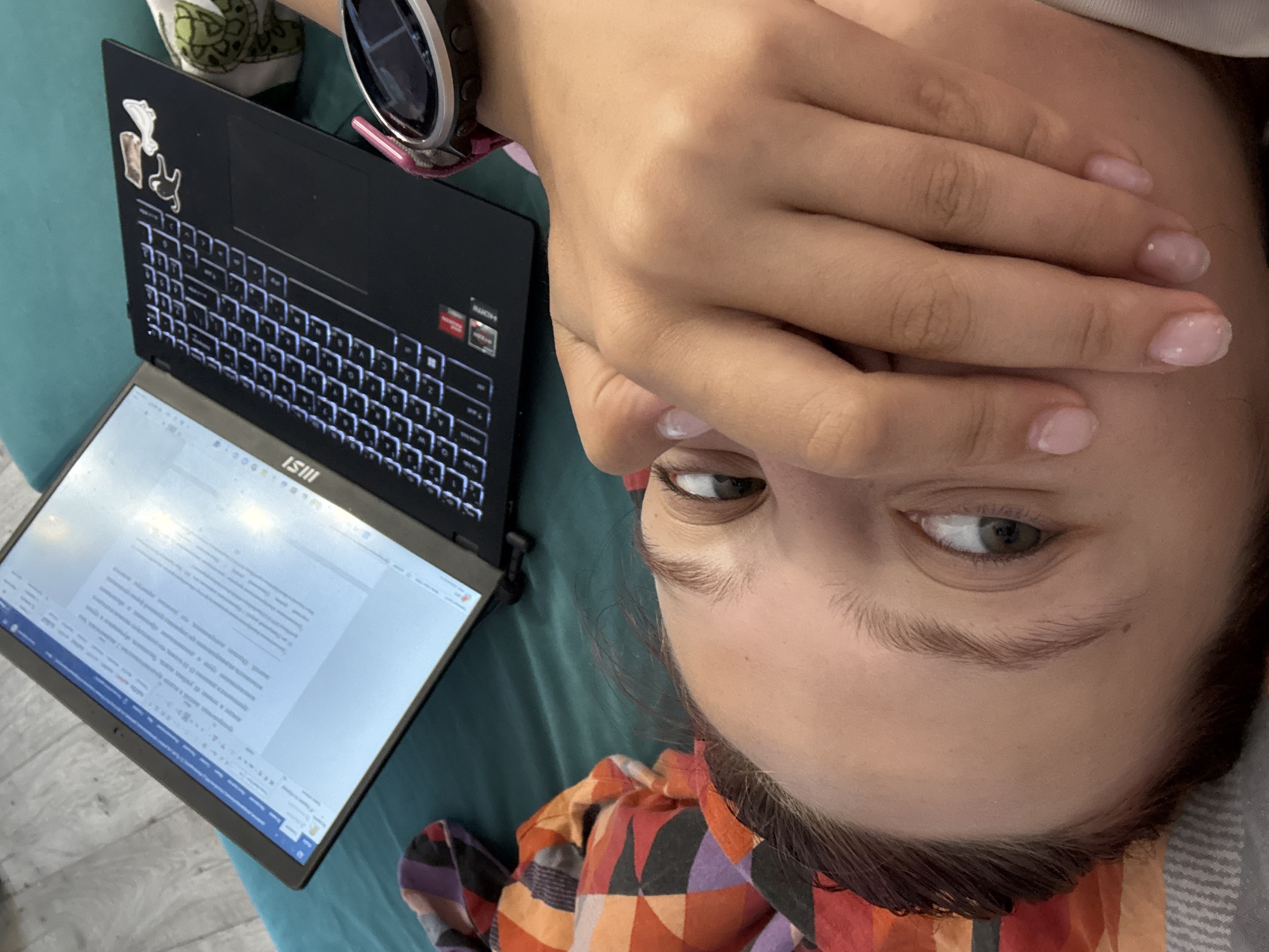 A person lies supine on a blue-green surface with a laptop positioned to their left. The laptop, an MSI brand, displays a Microsoft Word document filled with text. The person's right hand is raised, partially covering their face, revealing only their right eye. They wear a sports watch on their left wrist. The image suggests a casual or informal setting. There is no visual evidence in the image to indicate the location is Прикамский, Russia.