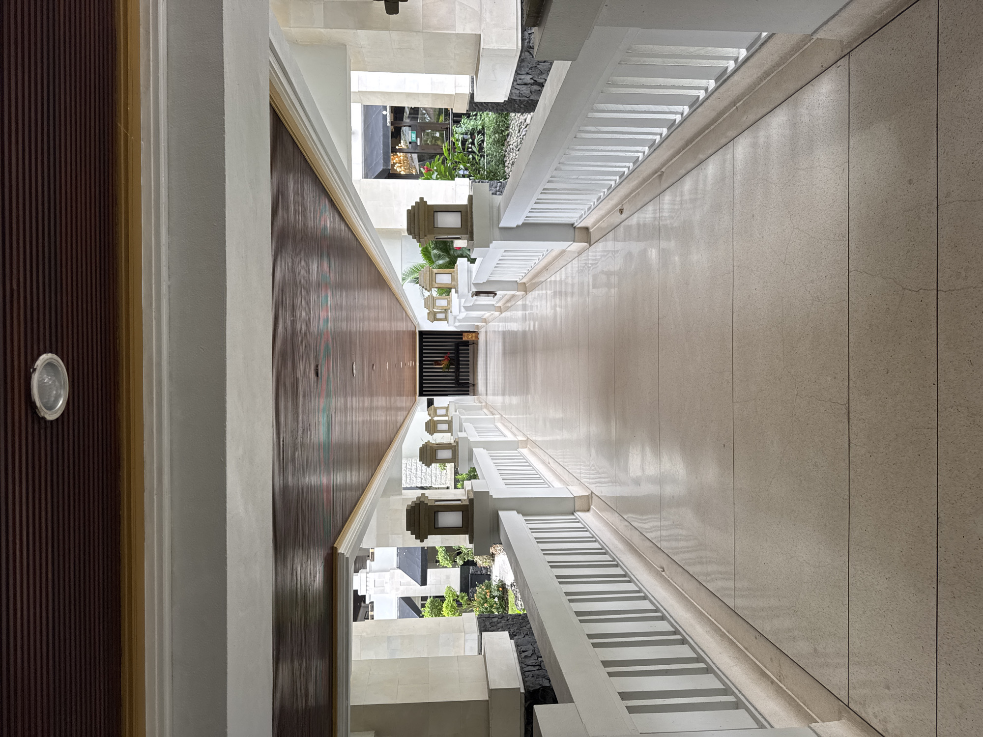 This is an interior shot of a hallway, likely located in a hotel or resort in Kuta Selatan, Indonesia. The hallway is oriented lengthwise, with a wooden ceiling on the left side and a light-colored tiled floor. On the right side of the hallway is a white railing, with a small garden visible beyond it. The walls are a light color, possibly concrete or tile. There are lantern-like light fixtures mounted on the wall. The hallway appears to be empty. The perspective is slightly distorted, giving a sense of depth to the space.