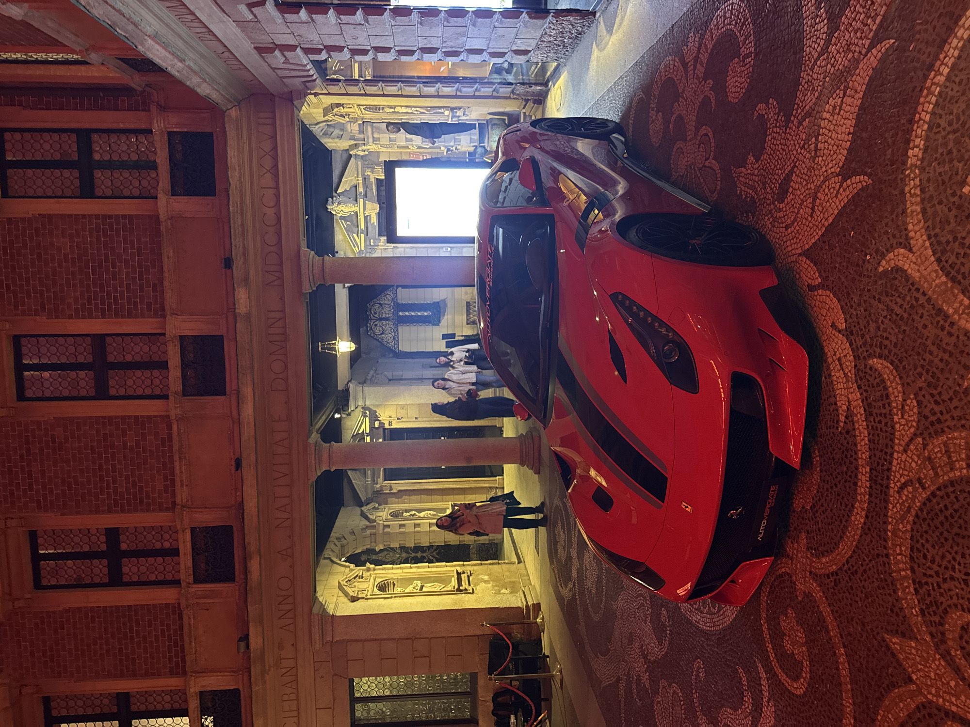 A red supercar, likely a Ferrari, is prominently displayed within an ornate architectural space, illuminated by warm artificial lighting, suggesting evening or night. The vehicle features "AUTOVERGIATE" on its front grille, along with a small Ferrari logo.
The setting appears to be a grand hall or covered courtyard of a historic European building, characterized by intricate details. The building's walls are constructed from brickwork, featuring multiple windows with circular grilles. Stately columns line a walkway, and decorative friezes adorn the structure. Sculpted statues are visible within niches along the walls, contributing to the classical aesthetic. The floor is a meticulously crafted mosaic, showcasing elaborate, flowing patterns in earthy tones.
Several individuals are present, observing the scene. One woman in a light-colored jacket stands near the supercar, facing the camera, while other people are visible in the background, near a brightly lit entrance. Red velvet ropes suggest a designated display area or controlled access for an event.
A significant detail is a Latin inscription carved into a frieze along the wall: "CIEBANT. ANNO A NATIVITATE. DOMINI. MDCCLXXVIII." This translates to "They ceased. In the year from the Nativity of the Lord 1778," indicating a historical date associated with the building or an event it commemorates. This, combined with the architectural style, strongly points to a historic location in Milan, Italy, as provided in the context. The overall atmosphere suggests an exclusive exhibition or event, harmonizing contemporary luxury with historical grandeur.