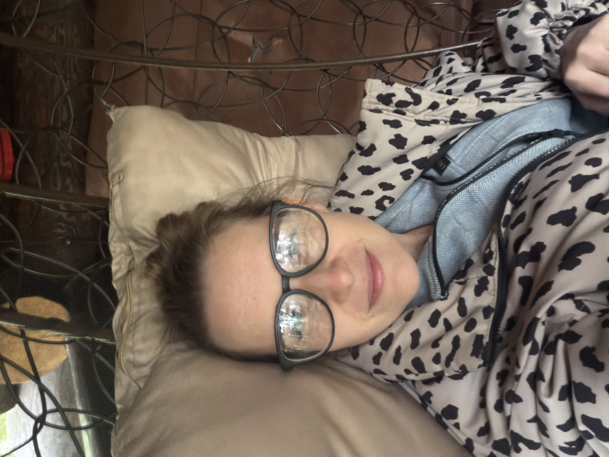 A woman wearing glasses is lying on a tan pillow in what appears to be a bed. She is wearing a jacket or coat with a black and tan animal print and a blue garment underneath. A metal bed frame with a decorative circular design is visible behind the pillow. The location, Zaplav'e, Russia, is not evident from the image.