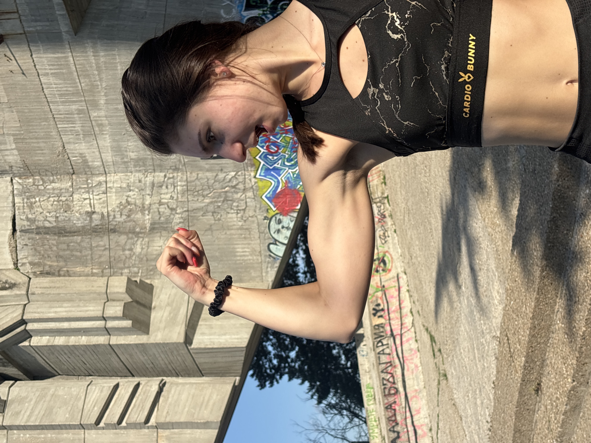 A light-skinned female is outdoors in Varna, Bulgaria. She is turned sideways, flexing her left arm and making a fist. She has dark hair pulled back in a ponytail. The woman is wearing a black crop top with a marble pattern and the words "CARDIO BUNNY" in gold letters. She is also wearing a black scrunchie on her left wrist. Graffiti-covered concrete structures are in the background, and a blue sky with trees is visible.