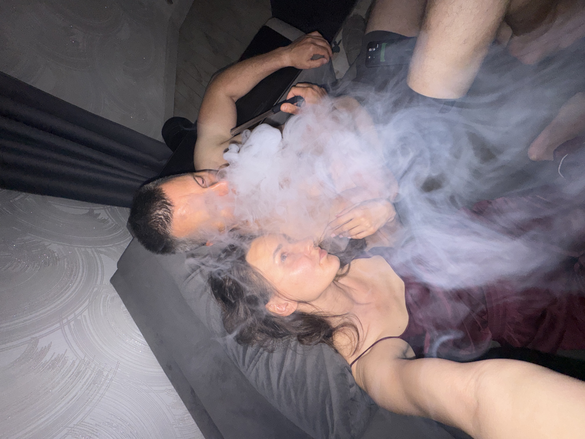 The image depicts two individuals, a male and a female, in a dimly lit indoor setting. The specific city of Sochi, Russia, requested in the prompt, cannot be visually confirmed from the image.
On the left, a female with dark, wavy hair is reclining on a dark gray pillow or cushion. She is wearing a dark red or burgundy spaghetti strap garment and is looking towards the right. Her left arm is visible, extended towards the bottom left corner.
On the right, a male is reclining, shirtless, and wearing dark shorts. He has short dark hair. He is actively exhaling a large, dense cloud of white vapor or smoke, which obscures part of his face and body, as well as the area between him and the female. He holds a clear glass tube with a dark handle, characteristic of a hookah or vaporizer mouthpiece, in his right hand. A black mobile phone with a green icon on its screen is visible on his lap or shorts.
The background features a light-colored wall with a textured, circular or swirling pattern, exhibiting subtle glitter or reflective particles. Dark gray curtains are visible on the far right. A dark gray upholstered headboard or section of a couch is positioned behind the female. A glimpse of light-colored flooring, possibly wood or laminate, is visible at the bottom right. The overall lighting is low, highlighting the vapor and the reflective elements in the room.