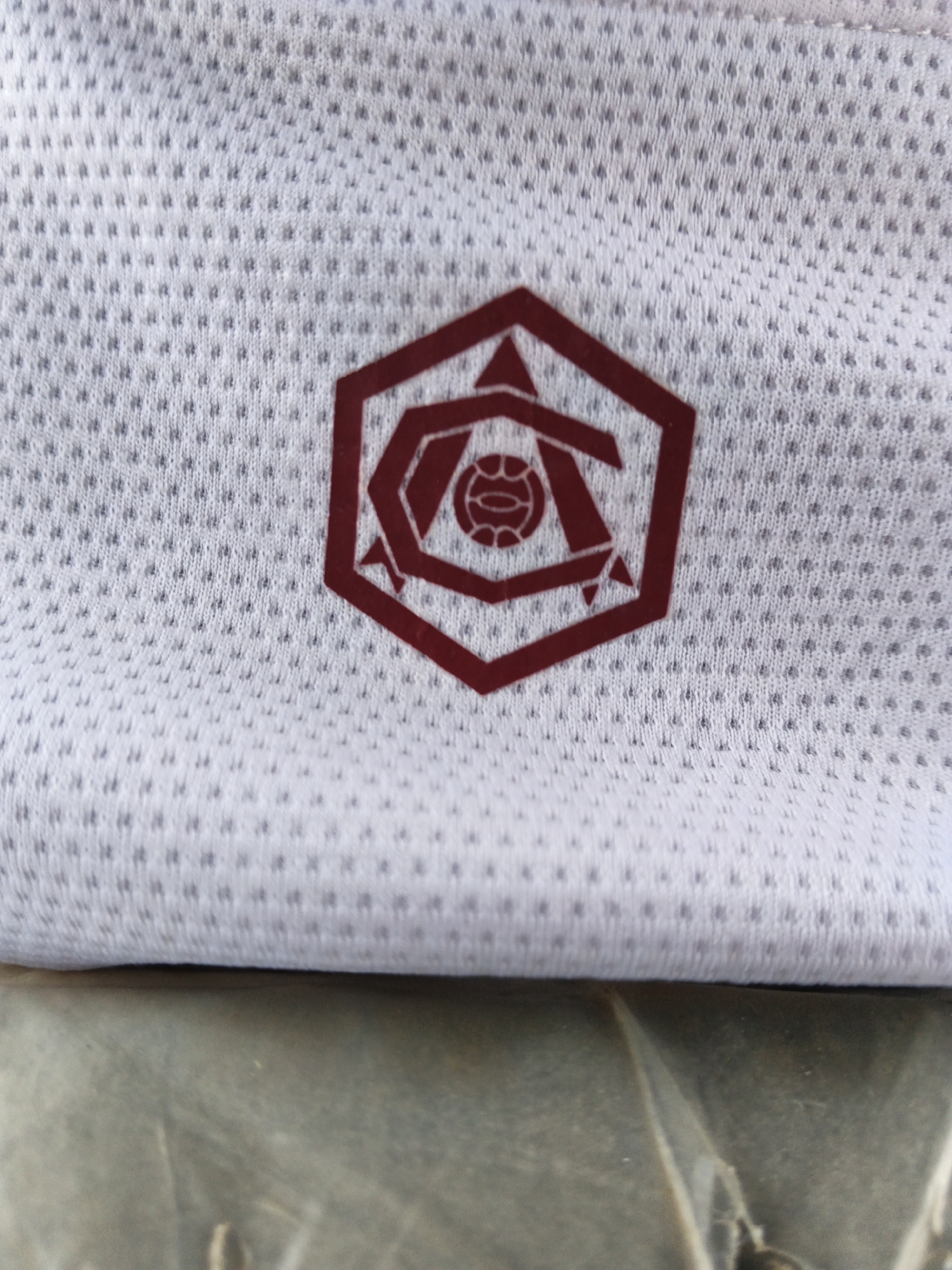The image shows a close-up of a white, textured fabric with a dark red hexagonal emblem. The emblem appears to be a logo, featuring abstract shapes and a circular element in the center that resembles a stylized soccer ball or planet. The fabric itself has a subtle dot pattern, characteristic of sportswear or athletic apparel. The lighting is even, suggesting an indoor setting or overcast conditions. The image focuses solely on the emblem and the fabric, with no other objects, people, or environmental details visible. No text is discernible within the image.