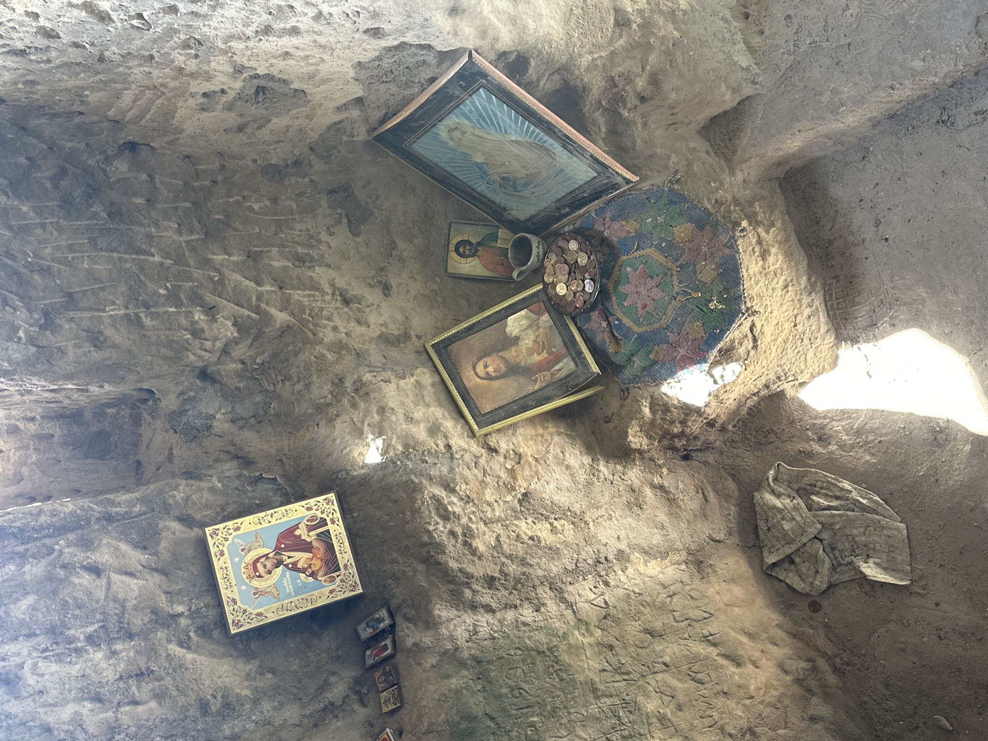 The image shows the interior of a cave or niche in Milanovo, Bulgaria. The walls are rough and appear to be stone or hardened earth. Attached to the walls are several religious icons and images in frames, of varying sizes and conditions. There is a plate or dish filled with coins placed near the icons, along with a ceramic mug. A woven rug or fabric is hung near the icons. The floor is covered with a cloth and has carved lettering or symbols on it. Sunlight enters from the right side of the frame, illuminating the interior.