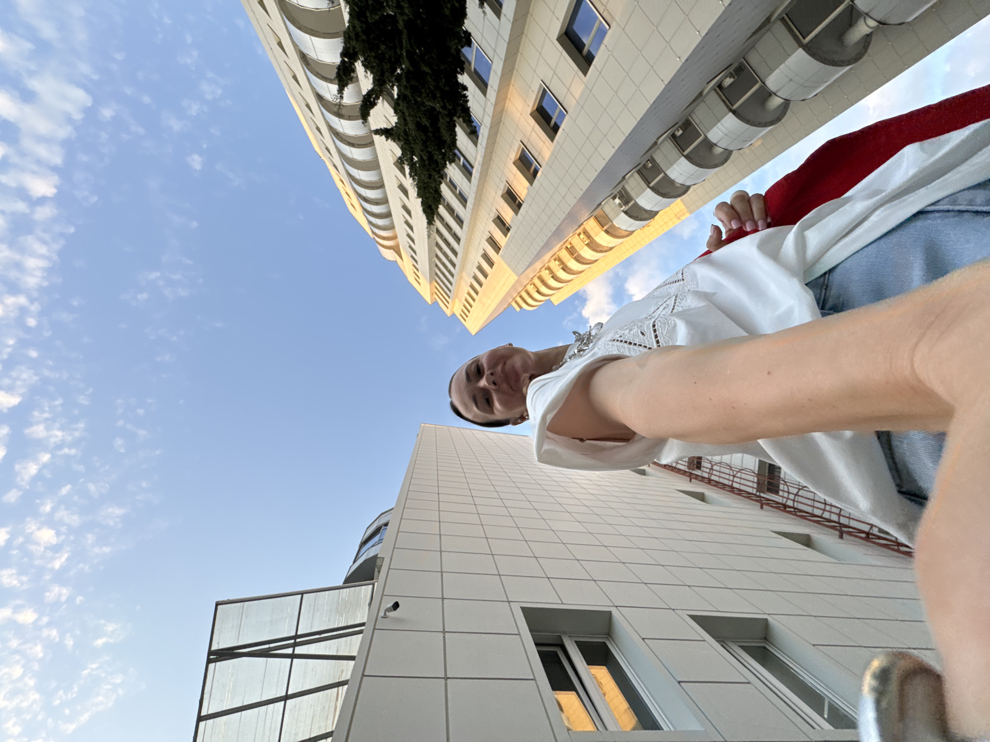 A low-angle, wide shot captures a person taking a selfie with a city skyline. The person, with dark hair pulled back, is wearing a white top and blue jeans. They are holding a red fabric with their right hand. In the background, two modern buildings dominate the scene, with the sky and scattered white clouds above. One building on the left is white with a tiled facade and a glass-covered section at the top. The building on the right is taller and features a curved design with multiple balconies, sunlight glinting off its facade. The scene appears to be set in an urban environment, possibly during daylight hours given the lighting.