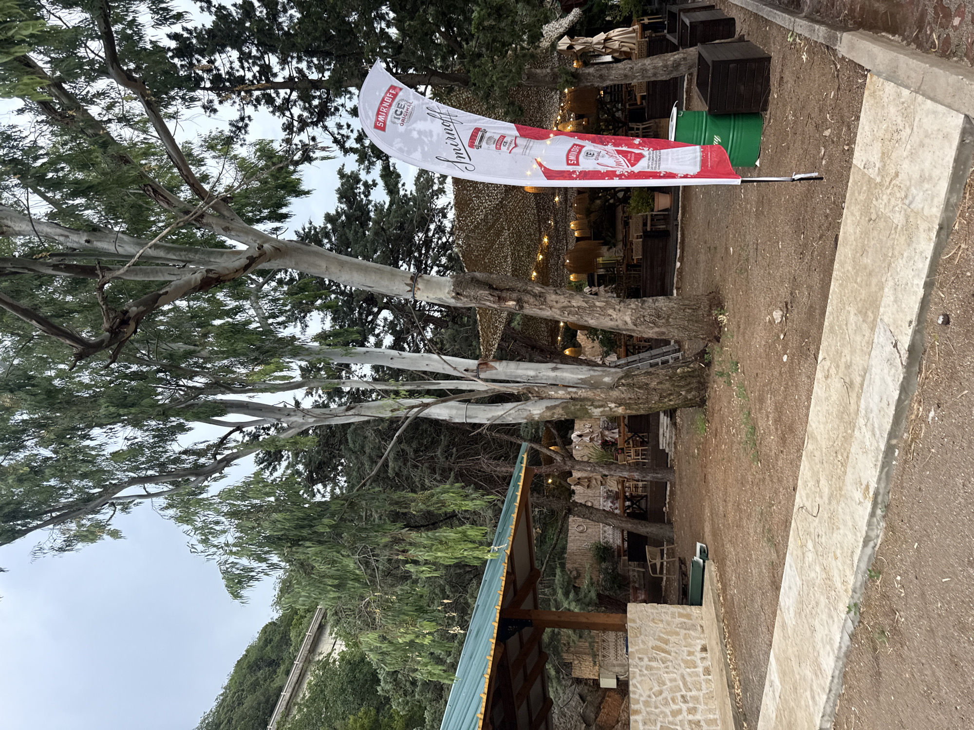 This image captures an outdoor scene at what appears to be a venue in Kamenovo, Montenegro. Dominating the frame is a large, white flag with red accents, prominently displaying the "Smirnoff ICE ORIGINAL" logo and branding. The flag is unfurled and appears to be made of a semi-transparent material, with a subtle mesh pattern visible. It is attached to a pole and positioned in front of a backdrop of trees and foliage.
In the background, glimpses of an outdoor seating area are visible, with tables, chairs, and umbrellas suggesting a restaurant or bar setting. String lights are also apparent, hinting at the possibility of an evening event. A large green barrel is situated on the ground near the flag. The ground is covered with a mix of dirt, gravel, and fallen leaves.
The scene is framed by the large, pale trunks and branches of several trees, which extend from the foreground into the upper portion of the image. Their leaves are a vibrant green, suggesting it is likely daytime. The sky above is overcast, indicating a cloudy day.
The overall impression is of a casual, outdoor gathering space, possibly associated with a brand promotion. There are no people clearly visible in the frame, but the setup implies a place for people to gather and relax. The presence of the Smirnoff flag strongly suggests an event or sponsorship activity related to the brand. The location cues, such as the stone structures and Mediterranean-style vegetation, are consistent with a coastal setting like Kamenovo.