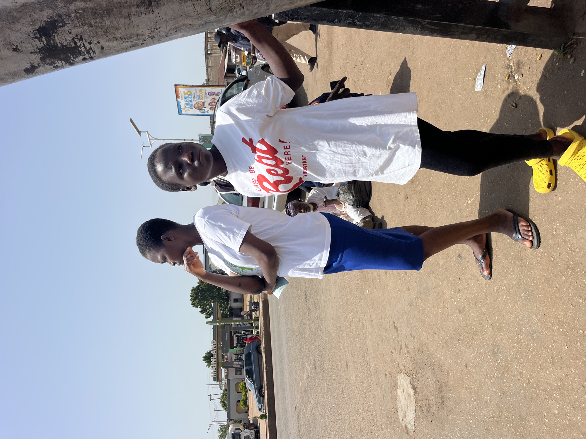 Two young Black females are standing on a dusty roadside in an urban environment. The female on the left is wearing a white t-shirt and a blue skirt, with black flip-flops. She has her left hand on her chin, looking to her left. The female on the right is wearing a white t-shirt with red lettering that reads "LASE BE R**t HERE! S IMPORTANT", black leggings, and bright yellow clogs. She is looking to her right. In the background, there is a gas station with a "MAINLAND" sign, several cars, and a billboard. A few other people are visible in the background, including a man in beige pants walking, and an older man sitting on the ground. The sky is clear and blue, suggesting it is a sunny day. The overall scene depicts a moment of pause or waiting on a street.