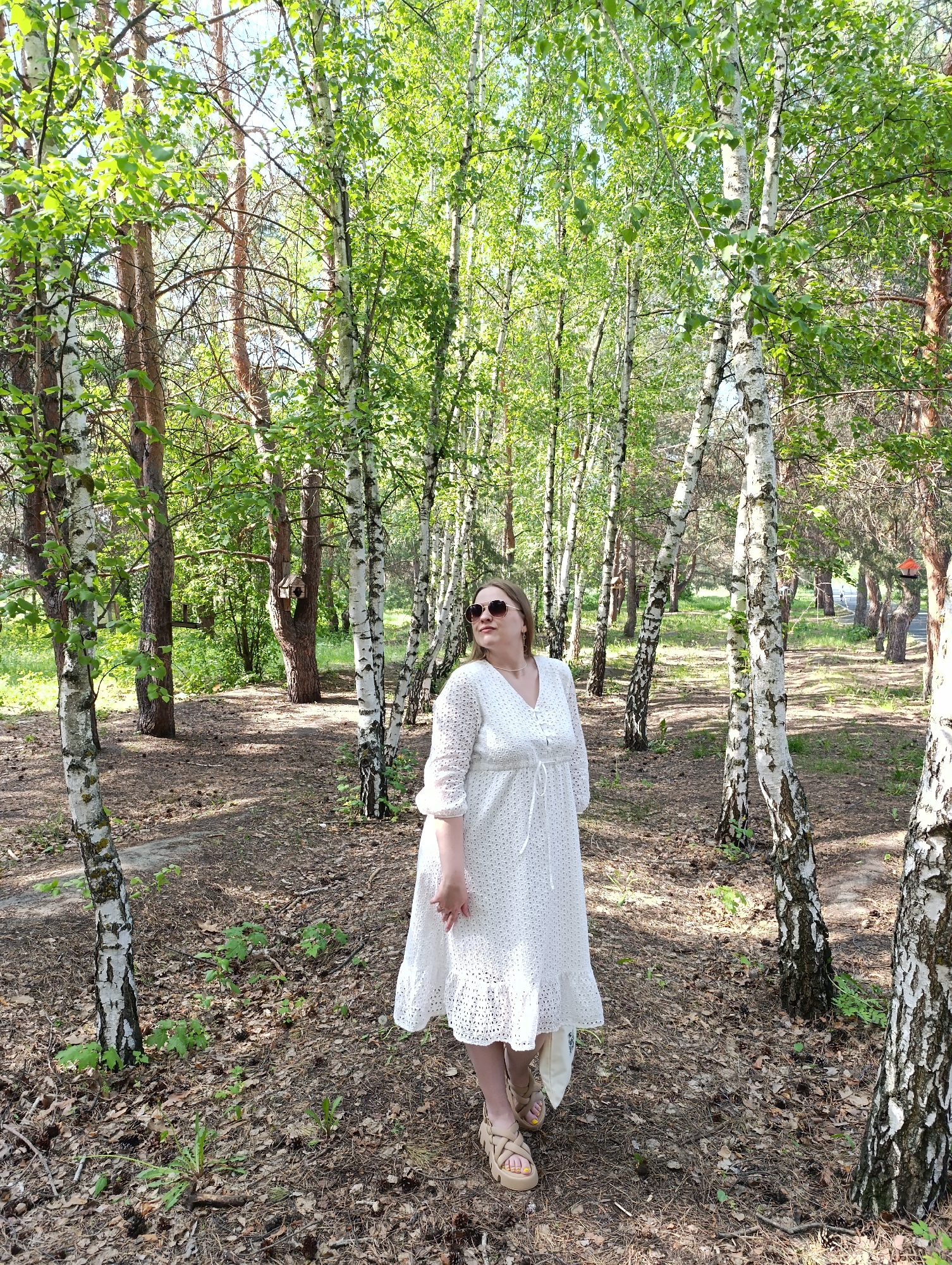A woman in a white dress and sunglasses stands in a forest of birch trees. The ground is covered in leaves and pine needles. The woman is looking to the left of the image. The trees are mostly bare, but some have green leaves. There is sunlight dappling through the trees.