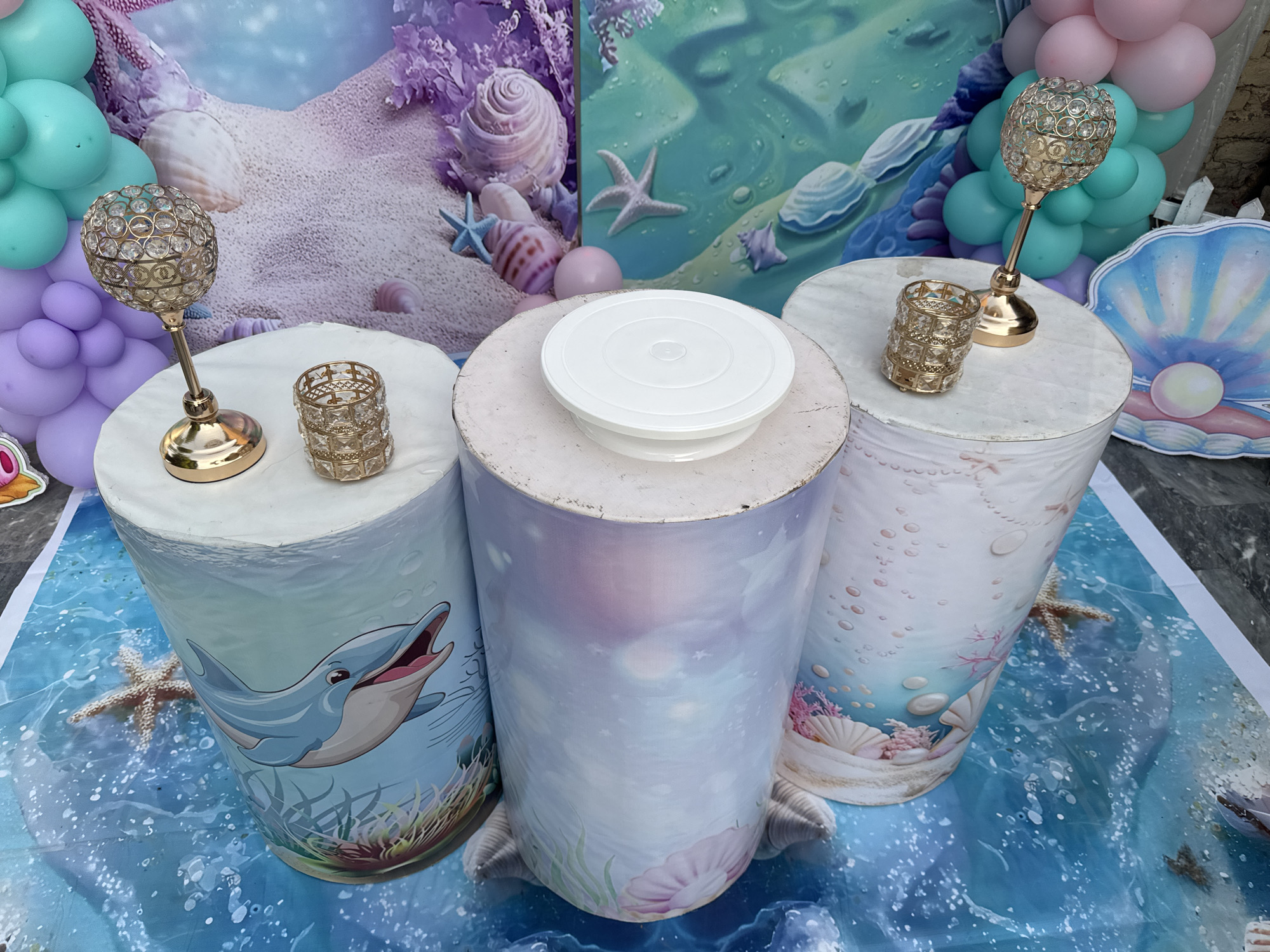The scene appears to be a party or event decorated with an underwater theme. There are three cylindrical platforms covered in themed graphics: one with a dolphin, one with an abstract underwater design, and one with marine life. The platforms are arranged on a blue and white surface depicting water. Each platform has decorative objects on top, including metallic candle holders and a cake turntable. The background includes balloons in pastel colors and a backdrop featuring underwater scenes with shells and starfish. The location is likely the Justice Colony area in Pakistan, given the query's reference to it, though there are no definitive visual indicators in the image to confirm this. There are no people visible in the frame.