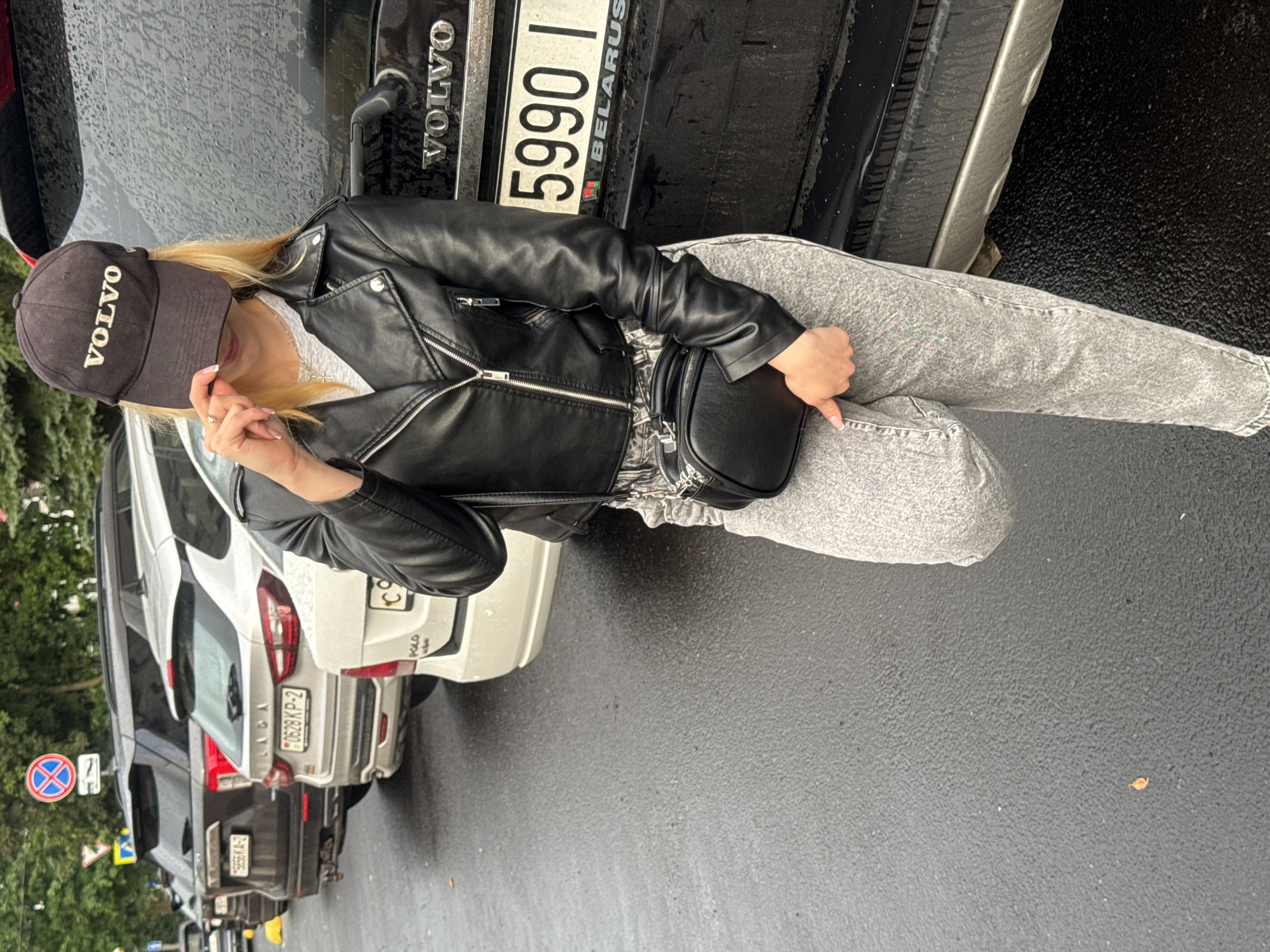 A woman is leaning against the back of a black Volvo car. She is wearing a black leather jacket, grey acid-wash jeans, and a black baseball cap with "VOLVO" written in white letters on the front. She has blonde hair and is wearing a white shirt underneath her jacket. She is also carrying a small black purse. The car has a license plate that reads "5990 I BELARUS." The ground is wet, indicating that it has recently rained. The scene appears to be a street or parking lot. There are other cars parked behind the Volvo, one of which is white and has a license plate that reads "0628KP-2." There are trees and traffic signs visible in the background. The time of day is unclear, but the weather is overcast.
