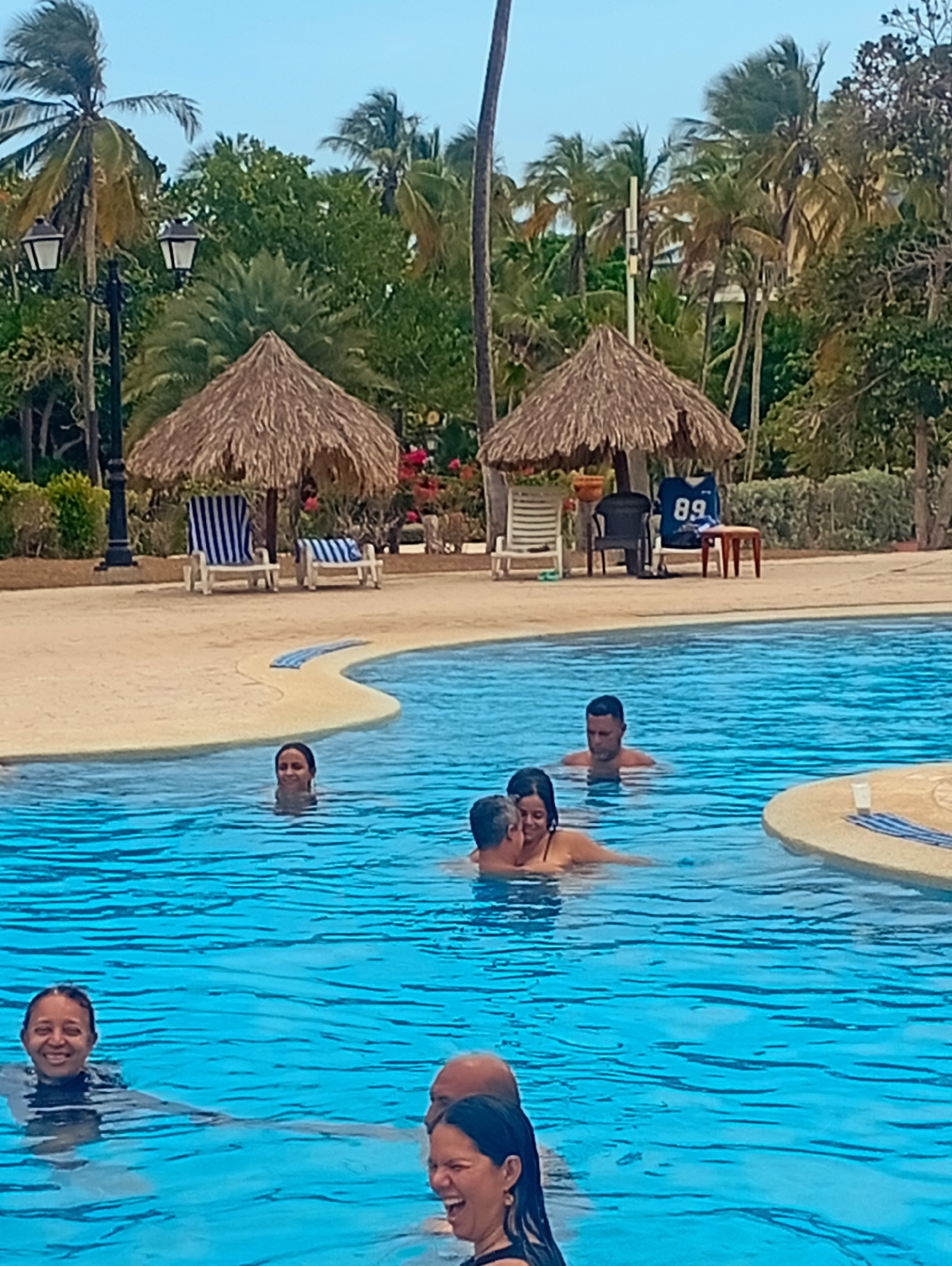 The image depicts a group of people enjoying themselves in a swimming pool. The scene is set in Playa El Agua, Venezuela, as evidenced by the tropical environment.
In the foreground, several individuals are partially submerged in the pool water, visible from the chest up. Some are smiling and interacting with each other.
In the background, there are two thatched-roof structures providing shade. Lounge chairs are positioned beneath these structures. Palm trees and other tropical vegetation are present.