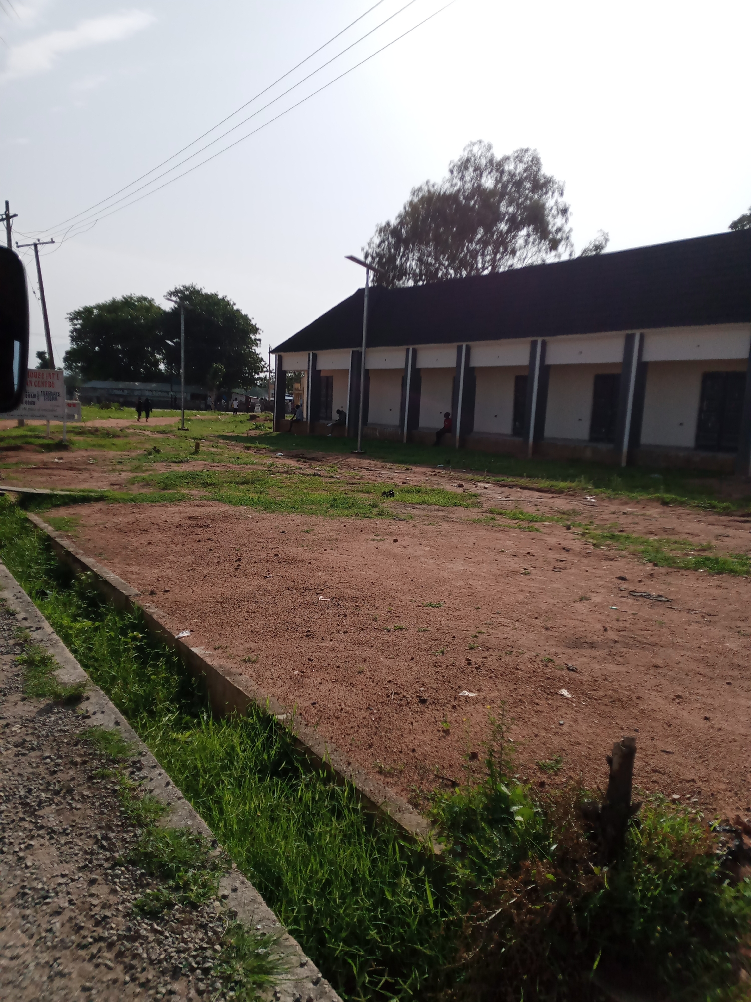 The image depicts an outdoor scene in Jalingo, Nigeria. In the background, a long, single-story building with a dark, sloped roof stands prominently. The building features a white facade with dark vertical columns and recessed sections that appear to be doorways or window frames. Several people are visible near the building; one individual is seated in the recessed area on the right, and a few others are walking in the middle distance. A large tree with dense foliage is situated behind the building.
The foreground is dominated by a large, open area covered in reddish-brown dirt. To the left, a concrete structure, possibly a drainage channel or curb, runs along the edge of the dirt area, with green grass growing in and around it. Gravel is scattered on the concrete. A utility pole with wires is visible on the left side of the frame. In the far left, a portion of a sign is visible with some text, including "HOUSE INT'L" and "AN CENTRE". The sky above is bright and hazy, suggesting daylight conditions. The overall impression is of a somewhat undeveloped or rural area on the outskirts of a city, likely a school or community center based on the building and signage.