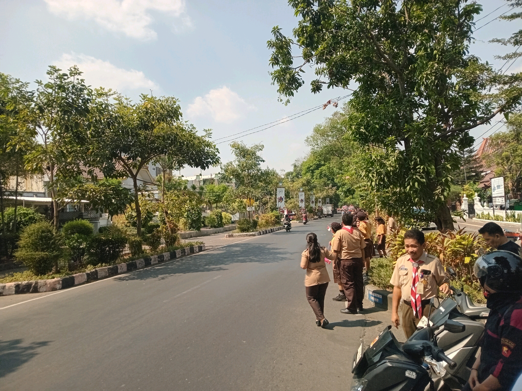 This image depicts a street scene in Surakarta, Indonesia, on a sunny day. In the foreground, several people are gathered near motorcycles, with some individuals wearing tan uniforms and red and white neckerchiefs, suggesting they are scouts. A man in a tan uniform with a neckerchief is speaking to someone out of frame, while other individuals in similar uniforms stand nearby, some holding flags. A woman in a tan uniform walks away from the viewer towards the street, and another person in a black helmet and jacket stands near a motorcycle.
The street itself is paved and appears to be a main thoroughfare, with a sidewalk lined with trees and bushes. Buildings are visible in the background, some with traditional Indonesian architecture featuring orange tiled roofs. There are also streetlights and power lines overhead. Traffic, including motorcycles and potentially other vehicles, can be seen in the distance. The overall atmosphere is one of an outdoor gathering or event.