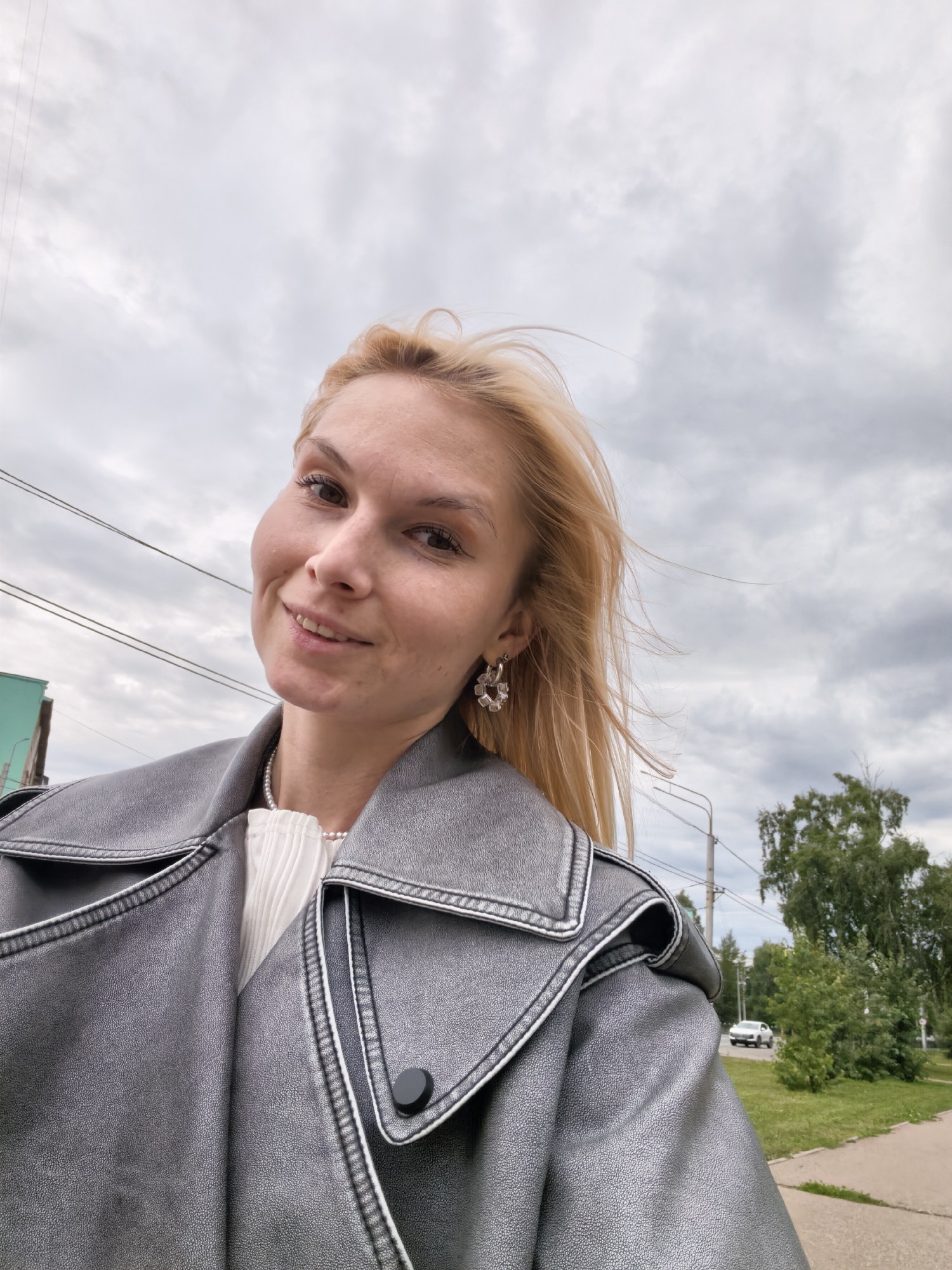 A blonde woman, wearing a white top and a silver leather jacket, smiles at the camera. She has on hoop earrings and a pearl necklace. The background shows a cloudy sky, trees, power lines, and a street with a white car. This image was taken in Sterlitamak, Russia.