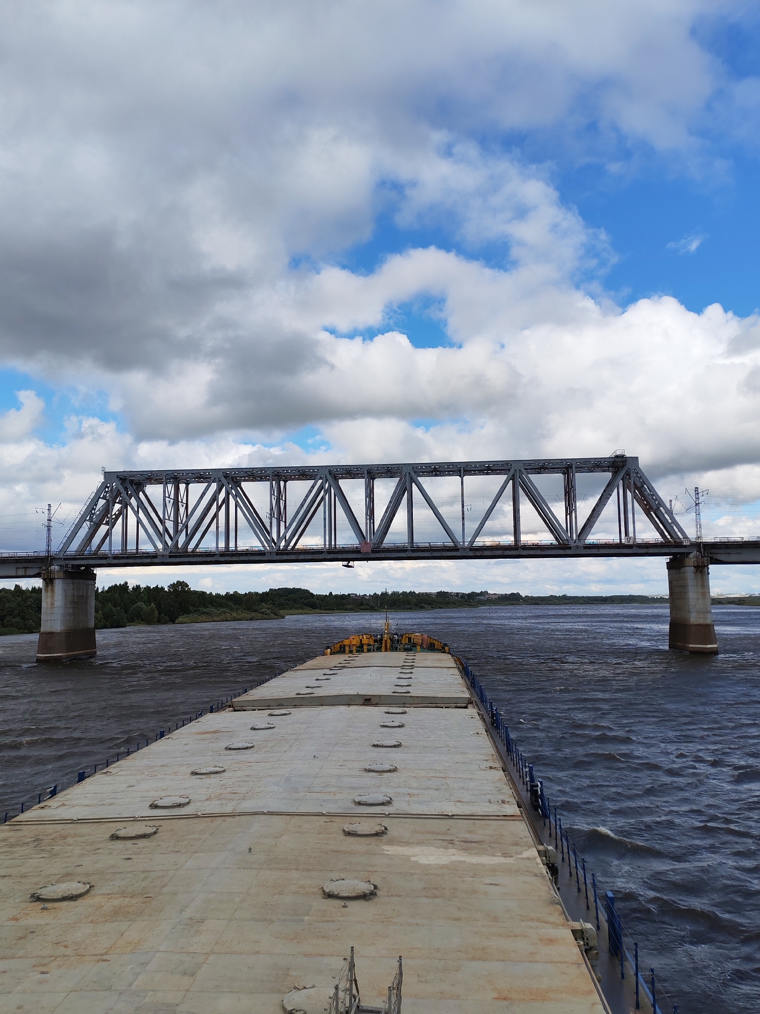 The image shows a wide, dark river with moderate waves under a cloudy sky with patches of blue. In the foreground, the flat deck of a barge is visible, with several circular hatches. The barge is moving forward towards a large metal truss railway bridge that spans the river. The bridge has concrete support pillars on either side of the river. The far bank of the river is lined with trees. The scene appears to be the Tarkanovo railway bridge over the Kama River, near Tarkanovo, Russia. There are no people visible in the image.