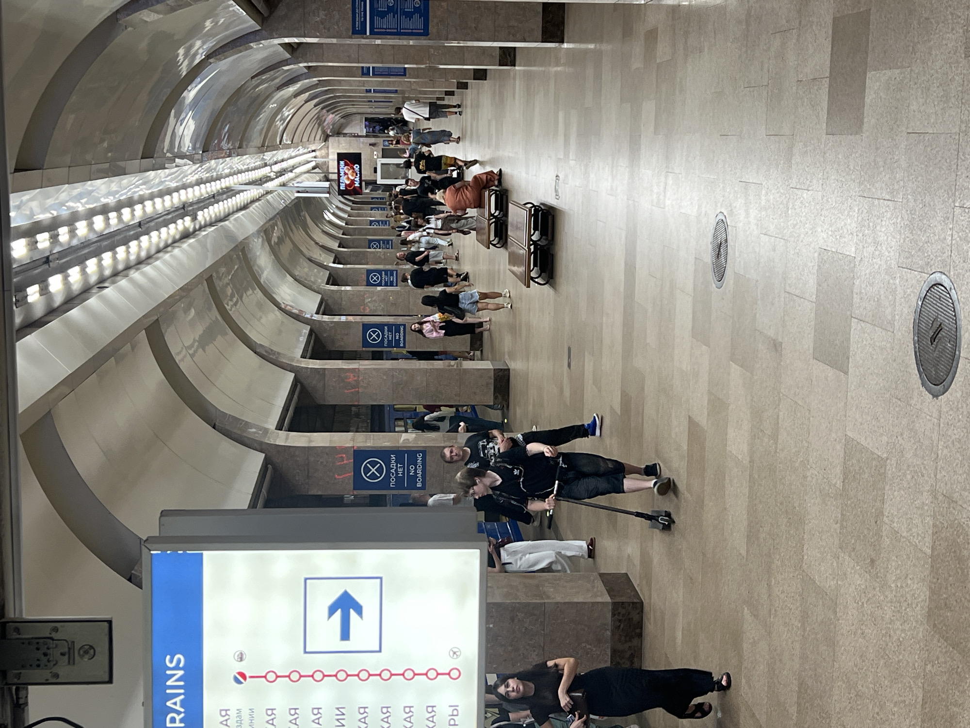 The image depicts the interior of a metro station in Nizhny Novgorod, Russia. The scene is characterized by a long, arched hallway lined with stone columns. People are seen walking in both directions along the tiled floor.
In the foreground, a woman in a black dress walks to the left. To her right, a young person is riding a kick scooter with a backpack. Further down the hallway, several other individuals are visible, some standing, some walking, and some sitting on benches.
On the left side of the image, a large illuminated sign displays "TRAINS" at the top, followed by a vertical list of Cyrillic text and a route map with circles and an arrow indicating direction. To the right of this sign, a blue sign with a circle and a diagonal line over a person symbol, along with the text "ПОСАДКИ НЕТ" and "NO BOARDING," is mounted on a column. Another similar sign is visible further down the hallway.
In the background, an illuminated advertisement for "ПЕЛЬМЕНИ" (pelmeni, a type of dumpling) with the word "МАСЛО" (oil) beneath it is displayed. The ceiling features a series of arches with integrated lighting, creating a bright and somewhat reflective environment. The overall impression is that of a functional, public transportation hub.