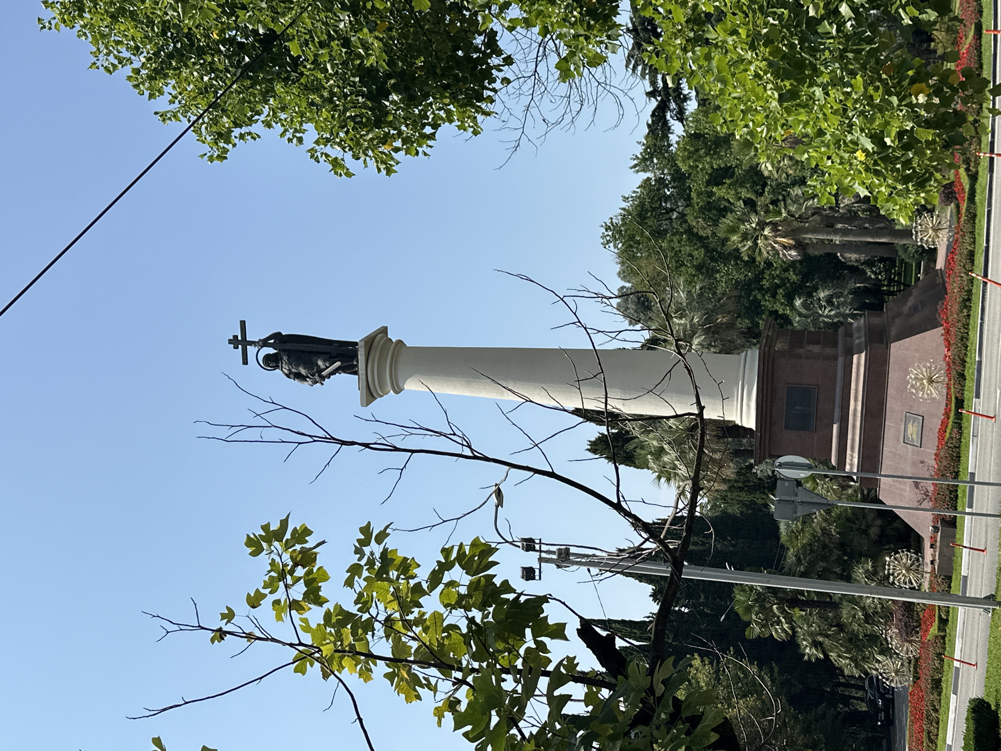 The image shows the monument to Archangel Michael in Sochi, Russia. It is a tall, white, cylindrical column topped with a bronze statue of Archangel Michael holding a cross and a sword. The monument is surrounded by lush green trees and a clear blue sky. In the foreground, there is a road with a traffic light and a bus stop. The monument is located in the center of the city, near the Black Sea coast.
