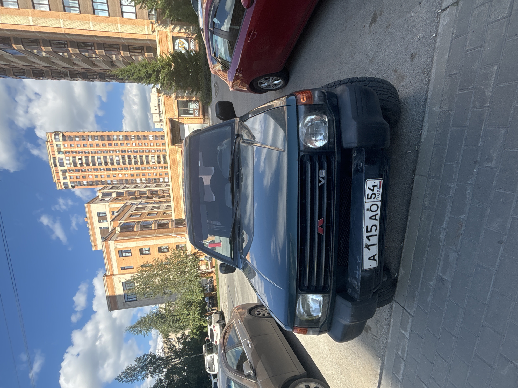 An outdoor, eye-level, close-up shot captures the front of a dark green Mitsubishi Pajero parked on a paved street in Novosibirsk, Russia. The vehicle's V6 emblem is visible on the grille. A license plate with the characters "A 115 AÖ 54 RUS" is affixed to the front bumper. In the background, multiple modern, multi-story buildings made of brick and glass are visible under a blue sky dotted with white clouds. To the left of the Pajero, a silver sedan is parked, and to the right, a red hatchback is present. The area appears to be a residential or commercial district within the city.