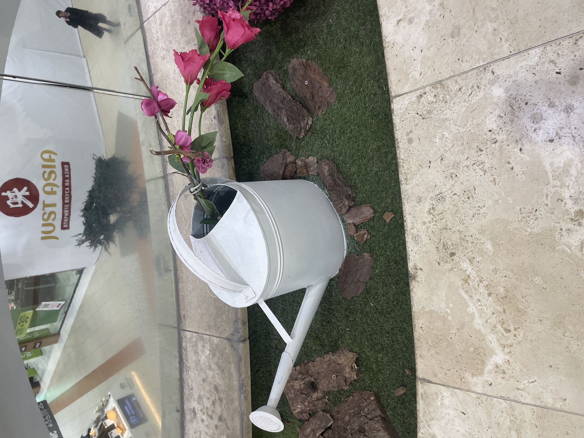 A white watering can filled with artificial pink flowers is positioned on a patch of artificial green turf. The turf is bordered by a light beige tiled surface and a glass window or wall. Scattered around the turf are pieces of dark brown tree bark. The reflection in the glass reveals a partial view of the interior of a building. Visible in the reflection is signage for "JUST ASIA". Also present are a few blurred figures in the reflection. This photo was taken in Sofia, Bulgaria.