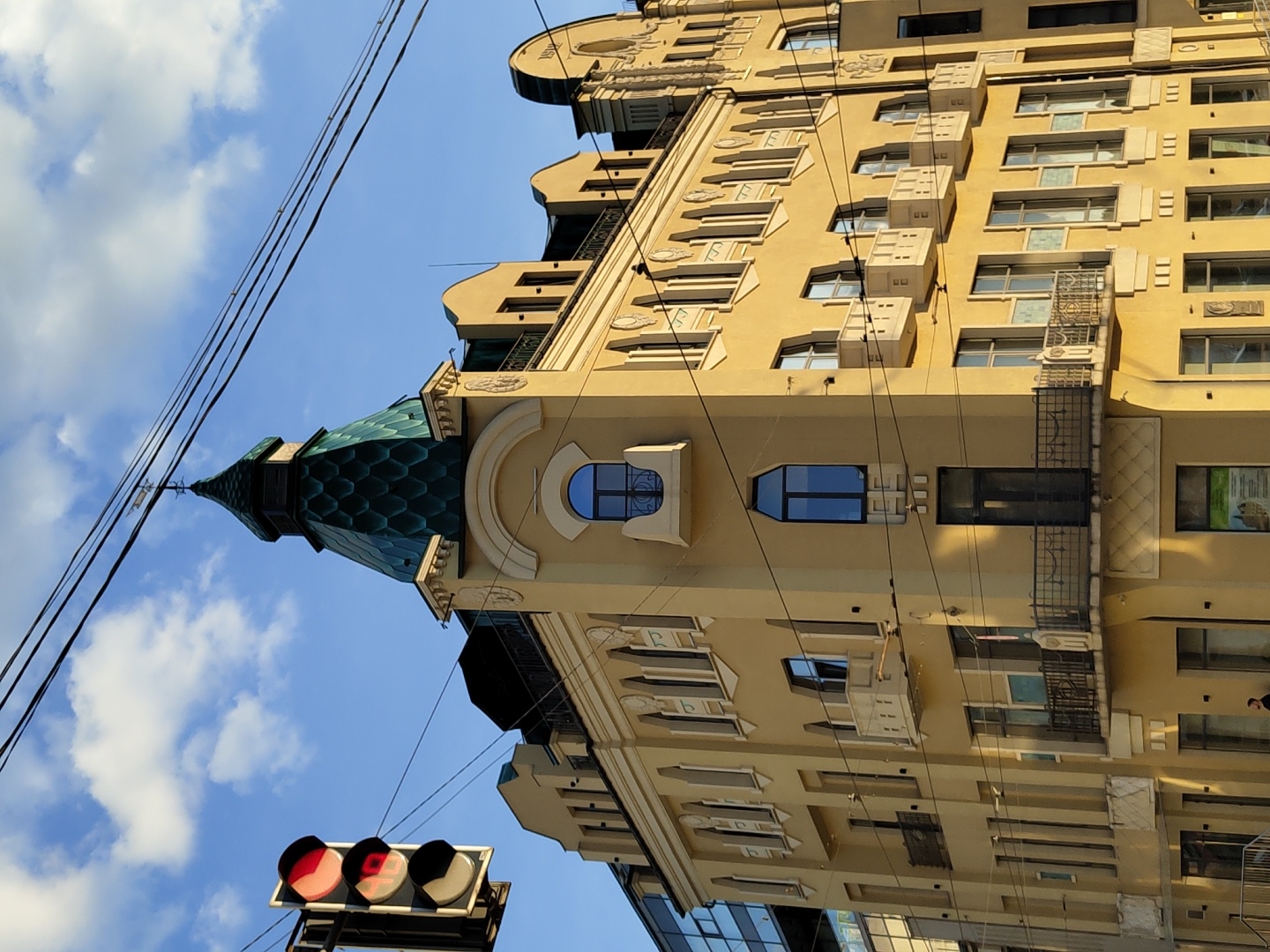 An upward-angled shot captures a multi-story building with a distinctive architectural style under a bright blue sky with scattered white clouds. The building's facade is a warm cream or yellow color, adorned with numerous windows of various shapes, including rectangular, arched, and circular designs.
A prominent feature is a tall, pointed dome or spire on the upper left side of the building, covered in a striking green, hexagonal scale pattern. The building also displays decorative architectural elements such as ornate moldings around windows, balconies with intricate railings, and textured wall panels. Several modern air conditioning units are affixed to the facade.
In the foreground, multiple dark utility lines stretch diagonally across the sky. To the lower left, a traffic light is visible, illuminated with a red signal. A digital countdown timer on the traffic light clearly displays the number "17". Parts of other buildings can be seen at the bottom of the frame.
The scene depicts an urban environment during daylight hours, indicated by the bright sun and clear sky. No people are visible, and the image captures a static architectural view. A blurry sign or poster with unreadable text and images is visible in one of the lower-right windows of the building.