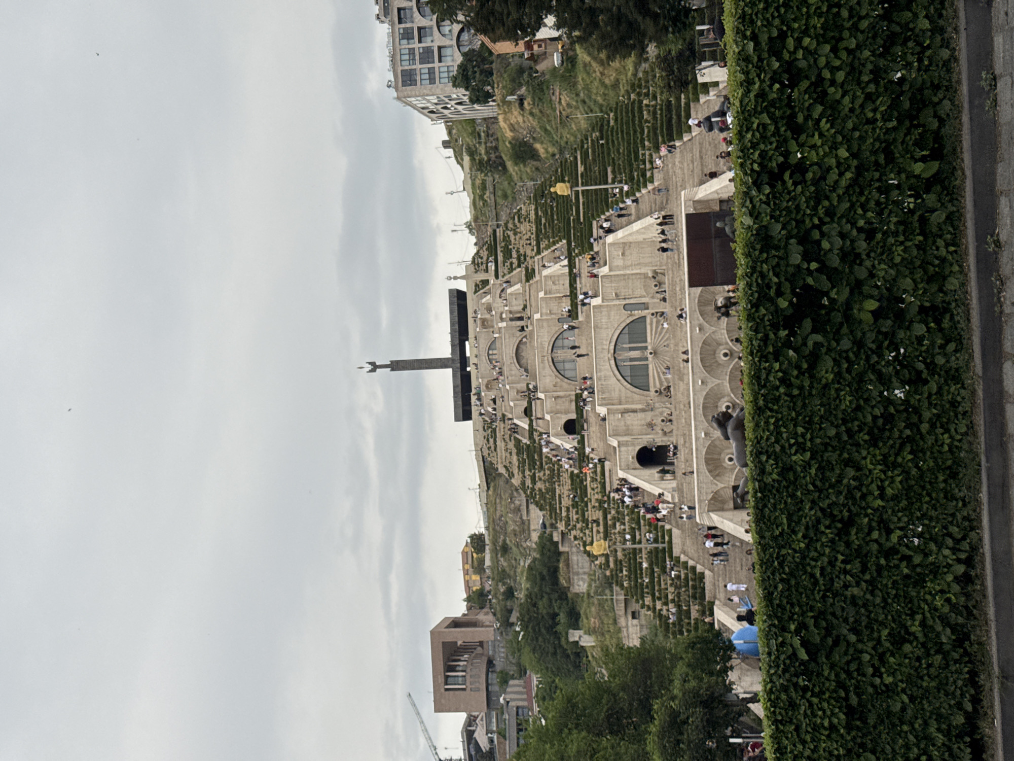 The image shows the Yerevan Cascade in Yerevan, Armenia, under a cloudy sky. The Cascade is a monumental stairway with multiple levels, decorated with fountains, sculptures, and flower beds. People are seen walking up and down the stairs. A tall obelisk-like structure stands at the top. Lush green bushes border the right side of the image, while buildings and greenery dot the background.