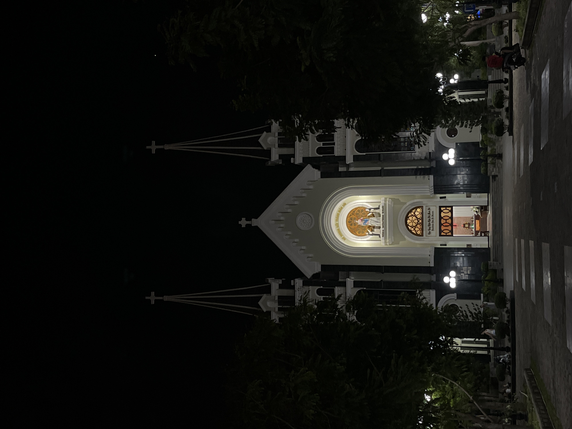 This image shows a night view of a church with two spires, illuminated against a dark sky. The church has a grand facade with a central arched entrance and a statue of a religious figure above. There are lights illuminating the entrance and the path leading to it. Trees and bushes surround the church, and a paved walkway with crosswalk markings is visible in the foreground. Several lampposts with multiple lights are placed around the grounds. In the lower right corner, a person is riding a scooter. In the background, there are glimpses of other trees and possibly more buildings, suggesting an urban or suburban setting. The overall atmosphere is serene and peaceful, typical of a religious site at night. While the prompt mentions the "city of Bac Nha Trang Ward, Vietnam," this specific location cannot be definitively confirmed from the visual information alone.