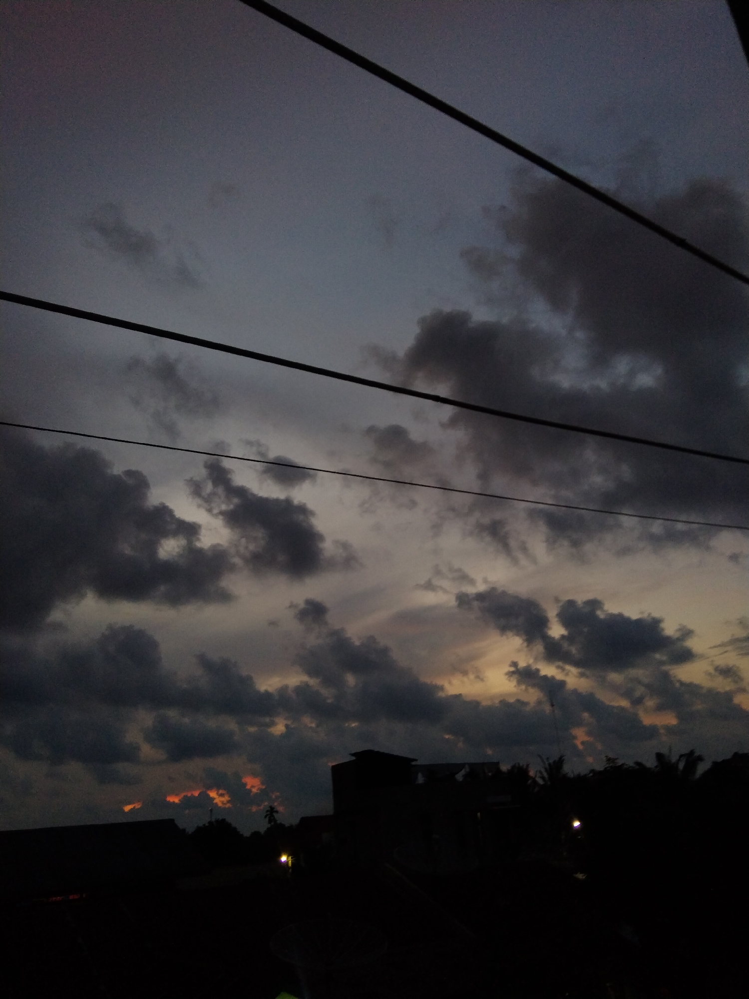 The image shows a twilight or dusk scene, likely in Bireuenmeunasahdayah, Indonesia, given the location tag. The sky is predominantly dark grey and filled with thick clouds, hinting at an overcast or stormy condition. Some areas display hints of orange or yellow near the horizon, suggesting a recent or upcoming sunset/sunrise.
Several dark silhouettes of buildings and trees are visible in the foreground, indicating an urban or semi-urban setting. Power lines are stretched across the top part of the frame. Two dimly lit light sources are visible among the silhouettes, possibly streetlights or lights from buildings. There are no people explicitly visible in the image.