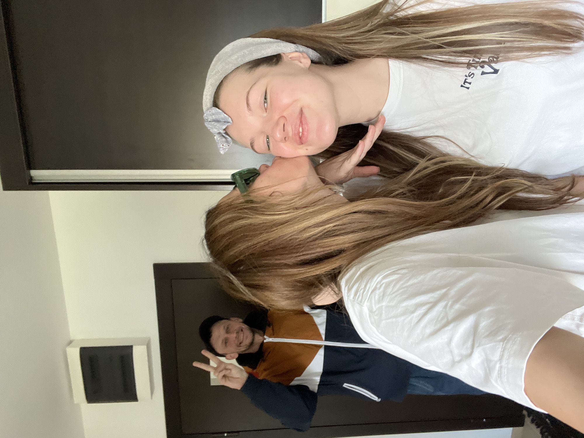 The image shows two young women and a man. The women are in the foreground. One woman is wearing a white shirt and a gray headband with a bow, and appears to be kissing the other woman on the cheek. The other woman is wearing green sunglasses and a white shirt. In the background, a man is visible, wearing a baseball cap and a blue, white, and brown jacket. He is making a peace sign with his fingers and smiling. The background features a door and a wall with a fixture on it.
There is no information in the image that indicates it was taken in the city of Vozdovac Urban Municipality, Serbia.