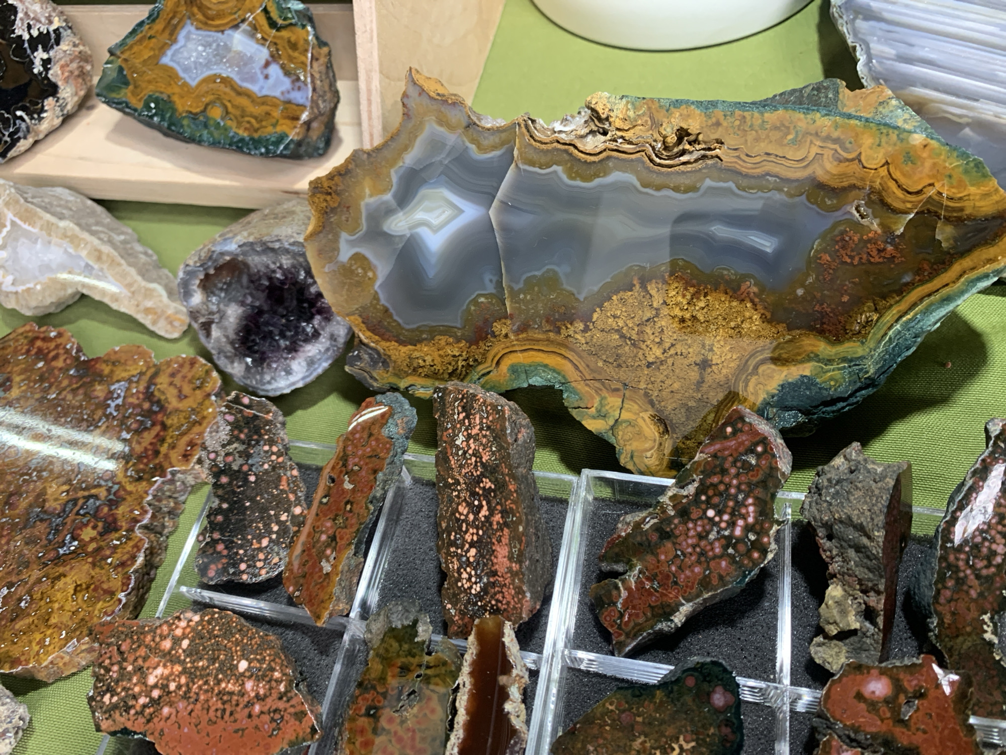The image shows a collection of polished and rough-cut gemstones and minerals displayed on a green surface, likely a table or countertop. In the center-right is a large, polished agate slice with concentric bands of gray, white, yellow, brown, and green. Below and to the left are several smaller pieces of what appears to be "poppy jasper" or a similar type of agate/jasper with reddish-brown coloring and orbicular patterns. To the left are also a geode with dark purple crystals, and a slice of banded agate with white and brown layers. The objects are arranged in a seemingly random pattern. The presence of mineral specimens suggests a rock shop, gem show, or private collection. There is no information available to confirm the location is Sofia, Bulgaria. There are no people present in the frame.