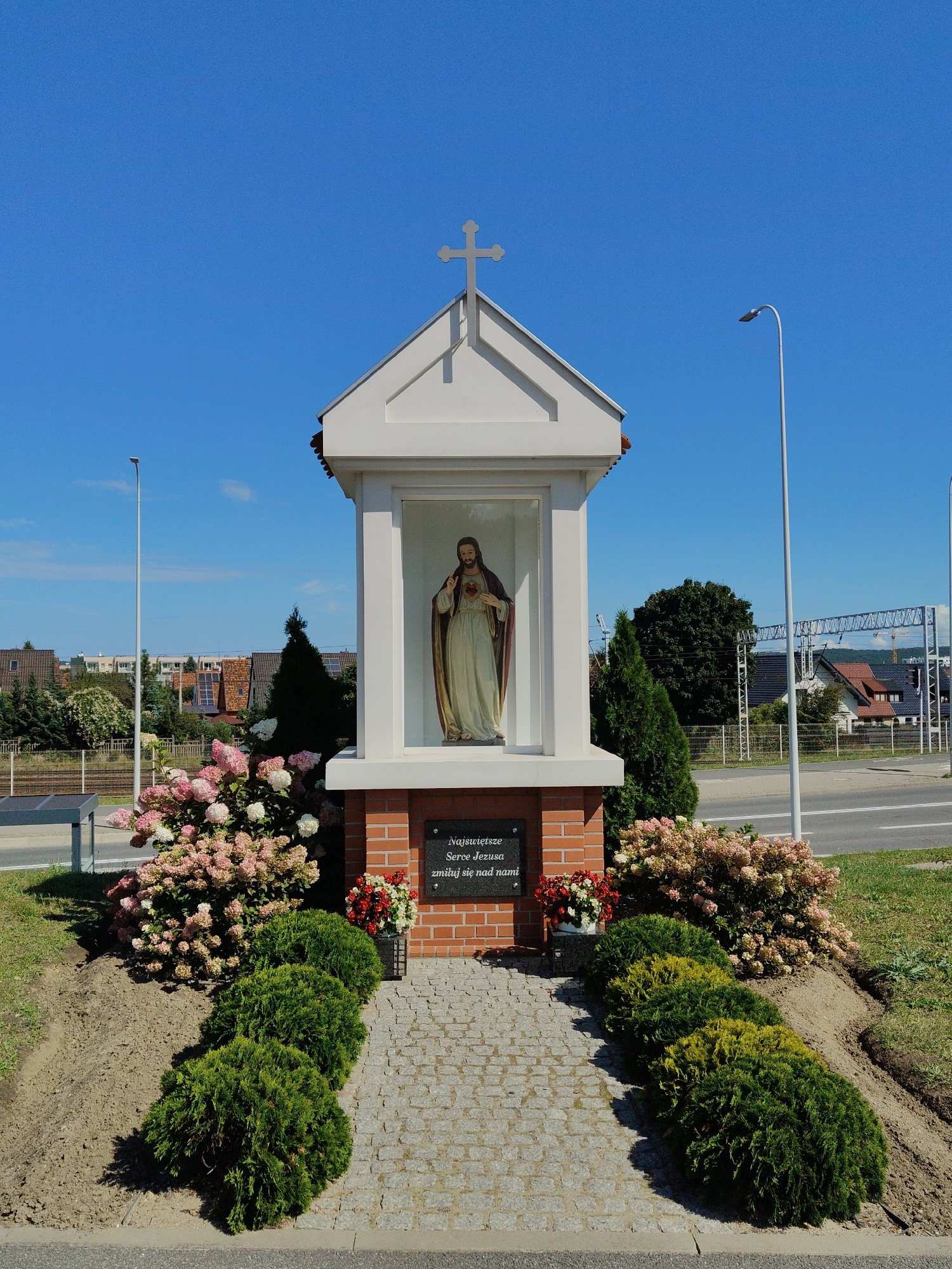 The image shows a small, white, shrine-like structure with a cross on its roof. Inside the shrine, a statue of Jesus Christ is visible, with his hand on his chest and a visible heart. The shrine is made of white material for the upper part and red brick for the lower part. A plaque with Polish text is attached to the brick base. The shrine is surrounded by a paved walkway and a landscaped garden with bushes and flowering plants. In the background, there are residential buildings, trees, a street, and a clear blue sky. This shrine is located in Rumia, Poland. The scene depicts an outdoor religious monument, with no people present. The environment suggests a sunny day.