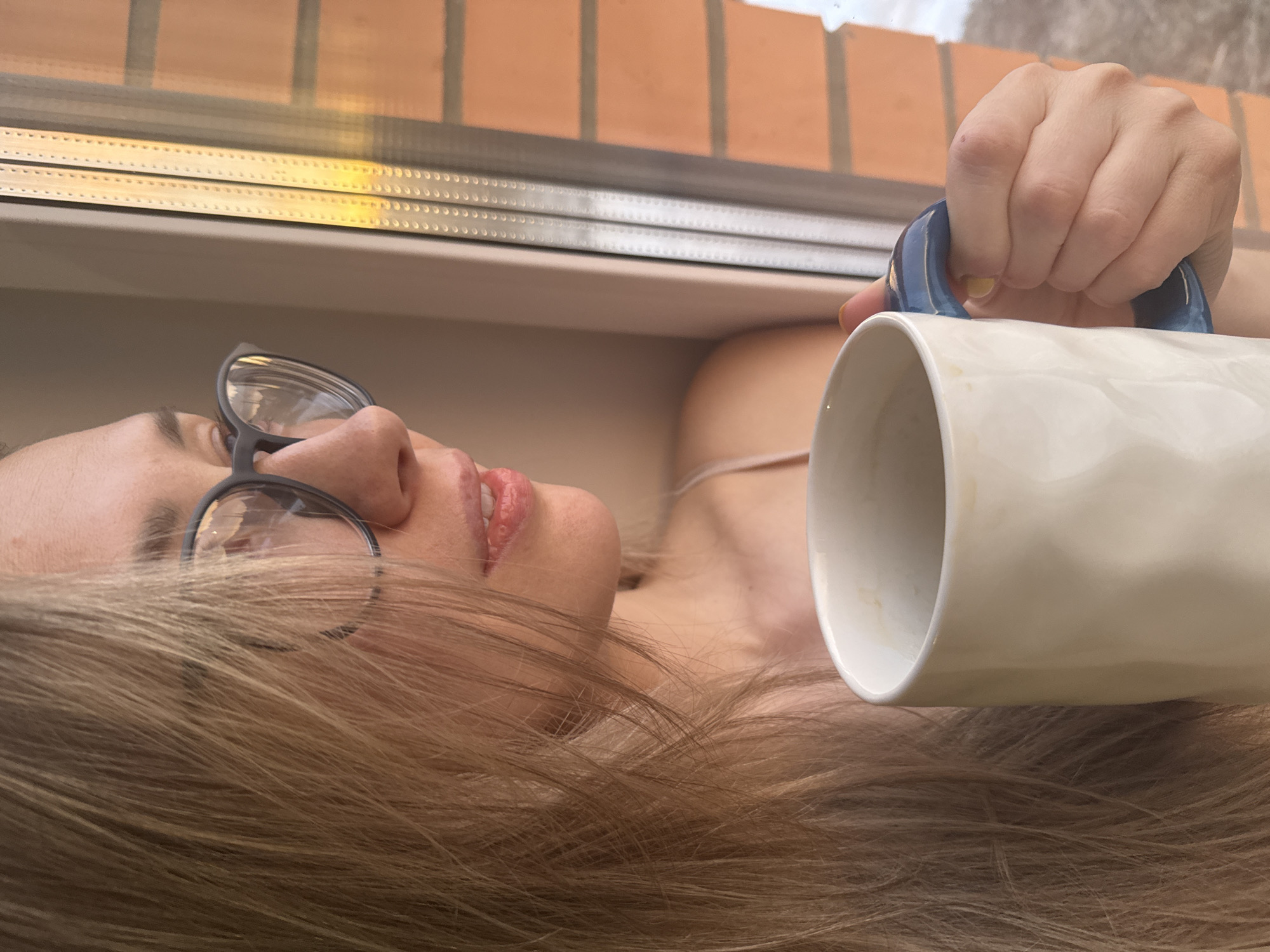 A fair-skinned woman with light blonde hair and glasses is lying down. She is holding a white mug with a blue handle. The location appears to be near a window, with a brick building visible outside. There is no definitive information to confirm that the location is in the city of Chaikovsky, Russia, but the architectural style of the building is consistent with that region.