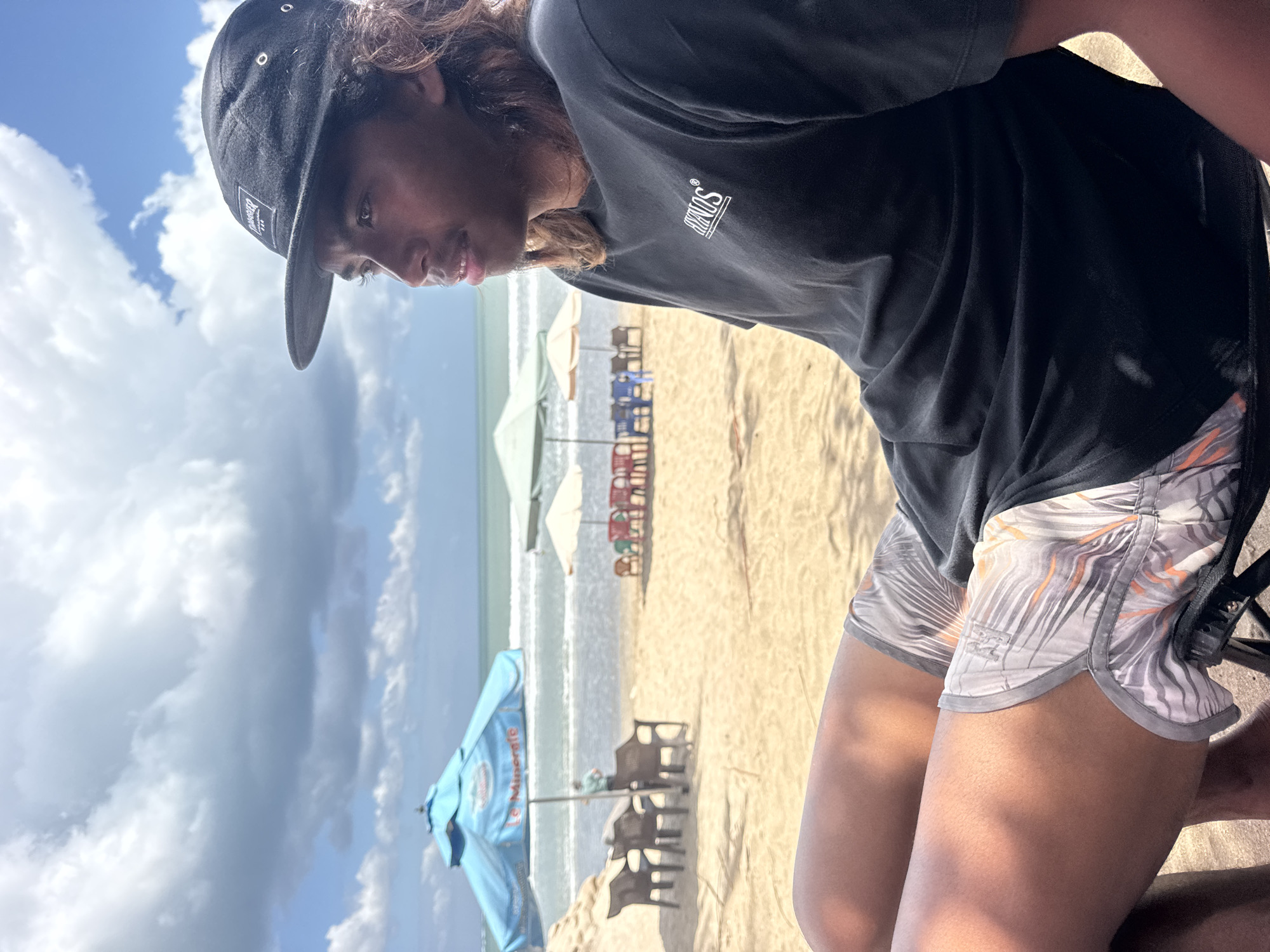 A young man with long, dark hair and a black baseball cap sits on a black folding chair on a sandy beach. He is wearing a black t-shirt with a white logo on the left chest and patterned swim trunks in shades of grey, white, and orange. He is looking to the right, out towards the ocean, with his legs crossed. In the background, there are several beach umbrellas, chairs, and tables. The ocean is visible, with small waves breaking on the shore. The sky is partly cloudy with blue patches. The scene appears to be in Kuta, Indonesia, a popular tourist destination known for its beaches.
