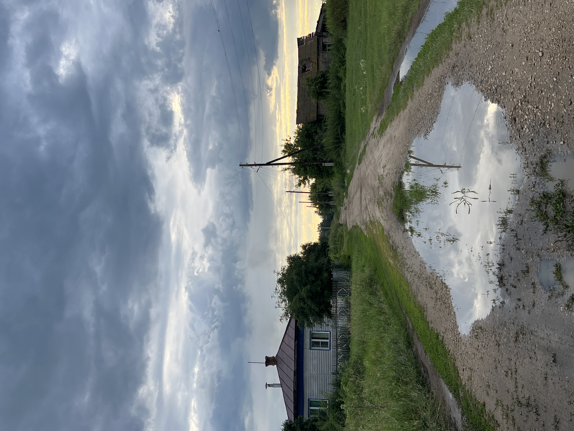 The image depicts a rural landscape in Pivkino, Russia, likely during the late afternoon or early evening, given the warm, soft light filtering through overcast skies. The scene is dominated by a dirt road that curves into the distance, flanked by overgrown grass and vegetation. A large puddle reflects the dramatic, cloudy sky and the surrounding environment, adding a sense of depth and mirroring.
On the left side of the road, a modest house with a dark-colored roof and light-colored siding is visible, bordered by a simple fence and dense greenery. Further in the distance, the silhouette of a partially ruined building, possibly an abandoned structure or barn, stands against the sky, hinting at the passage of time and decay. Utility poles with power lines traverse the sky, indicating the presence of infrastructure even in this seemingly remote setting.
The weather appears to be partly cloudy, with heavy, dark clouds in the upper left portion of the frame gradually giving way to brighter, more illuminated patches of sky near the horizon. This contrast suggests a dynamic weather system, perhaps a storm passing or approaching, which contributes to the somber yet beautiful mood of the photograph. The overall impression is one of quiet rural life, touched by elements of nature's power and the signs of human habitation, both present and past. There is no visible text in the image.