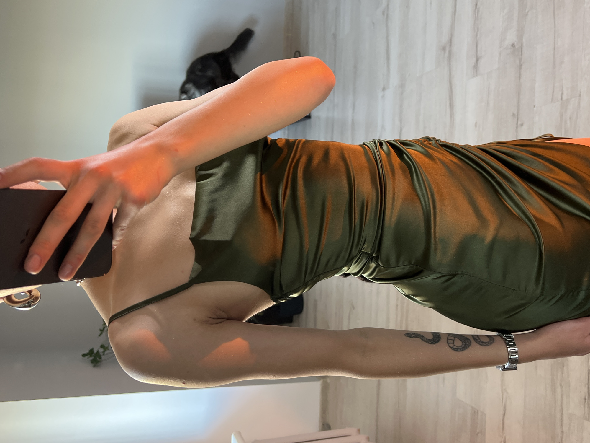A woman is taking a selfie in what appears to be an indoor setting. She is wearing a green satin dress. Her left arm is visible, featuring a tattoo of a snake and a metal watch. She is holding a dark-colored smartphone in her left hand, presumably to take the photo. The background shows a room with light-colored walls and flooring that resembles wood. A black cat is partially visible in the background. There is nothing in the image to suggest a specific location within the city of Ternopil, Ukraine.