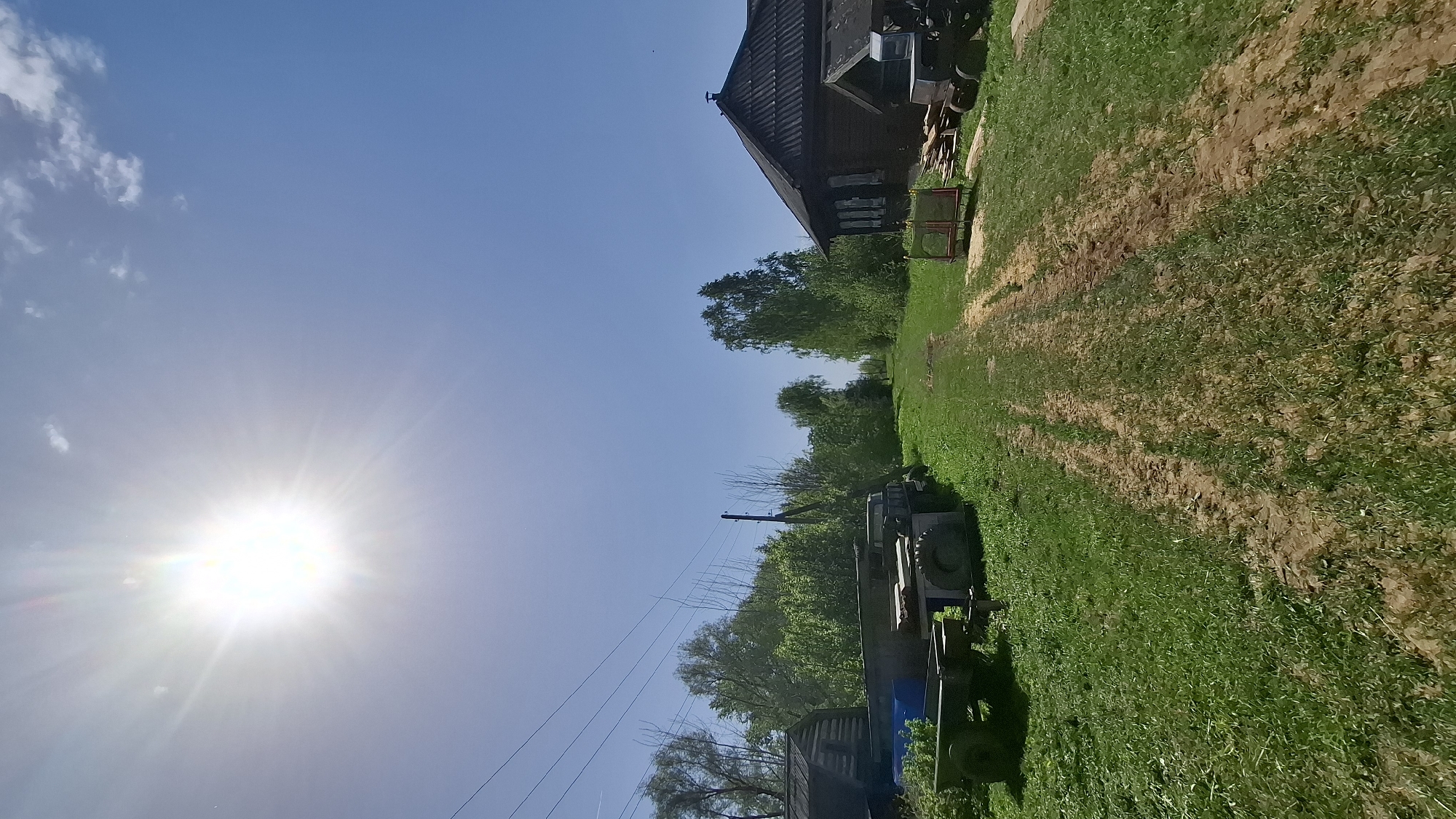 A daytime landscape photograph taken in Sheshurga, Russia, shows a rural scene. The sun is bright in the upper left, shining across a blue sky with some clouds. To the right, a dirt path with tire tracks runs from foreground to background, bordered by green grass. An old wooden building is visible at the top right. A tractor is parked near the center-right of the image. Trees are visible in the mid-ground. Power lines run across the lower left of the image.