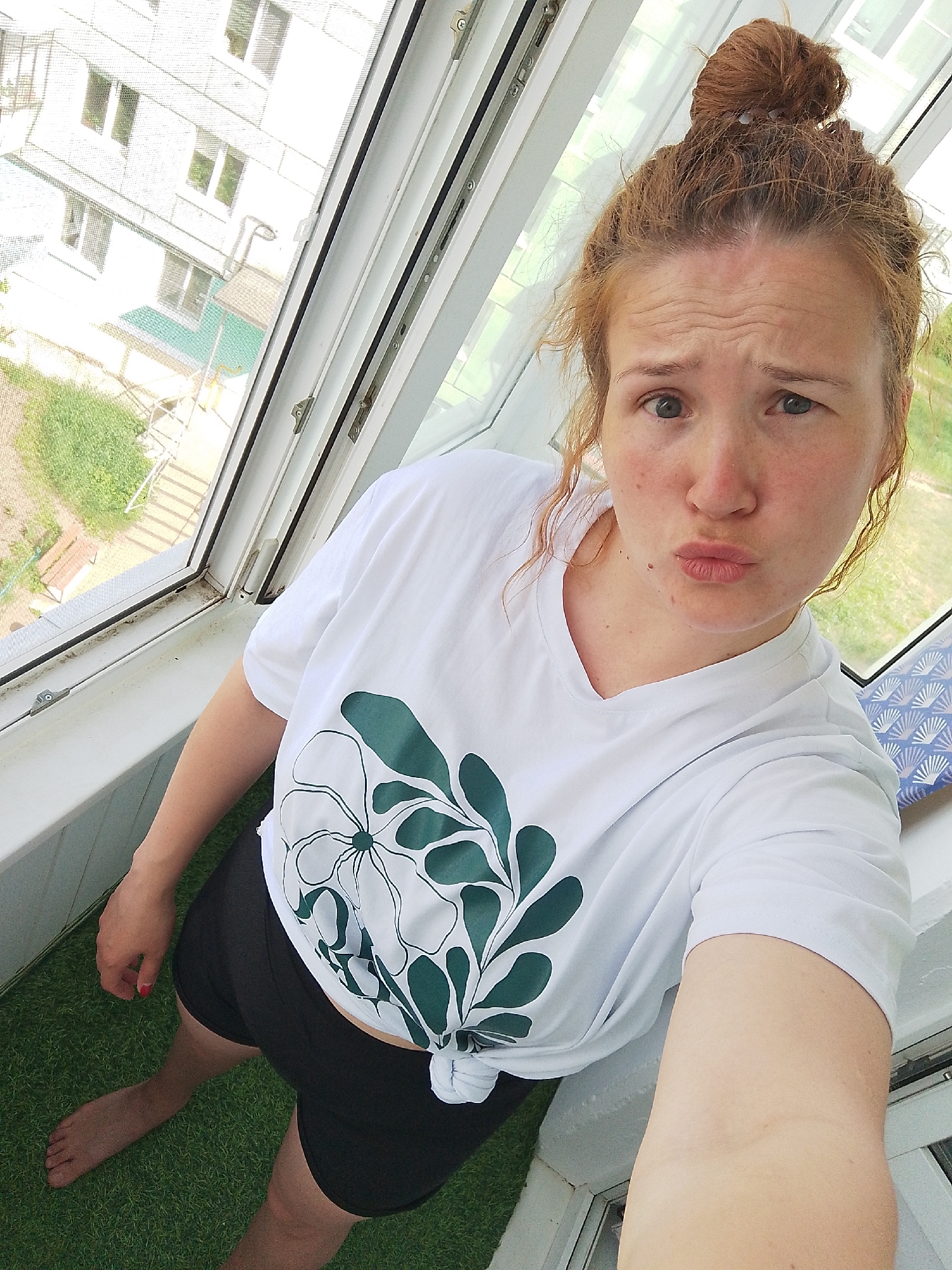 A medium-shot, high-angle photograph depicts a fair-skinned woman with red hair styled in a messy bun standing on what appears to be a balcony or enclosed porch. The woman is wearing a white t-shirt with a large green and white floral design on the front, which is knotted at the waist. She is also wearing black shorts and is barefoot on what seems to be artificial grass. Her facial expression includes slightly pursed lips and a furrowed brow.
Behind the woman, a large window frame is visible, revealing a view of a building with multiple windows and some greenery below. The environment suggests a residential area. The overall lighting is bright and natural. The setting appears to be indoors/semi-outdoors in Unknown, Unknown.