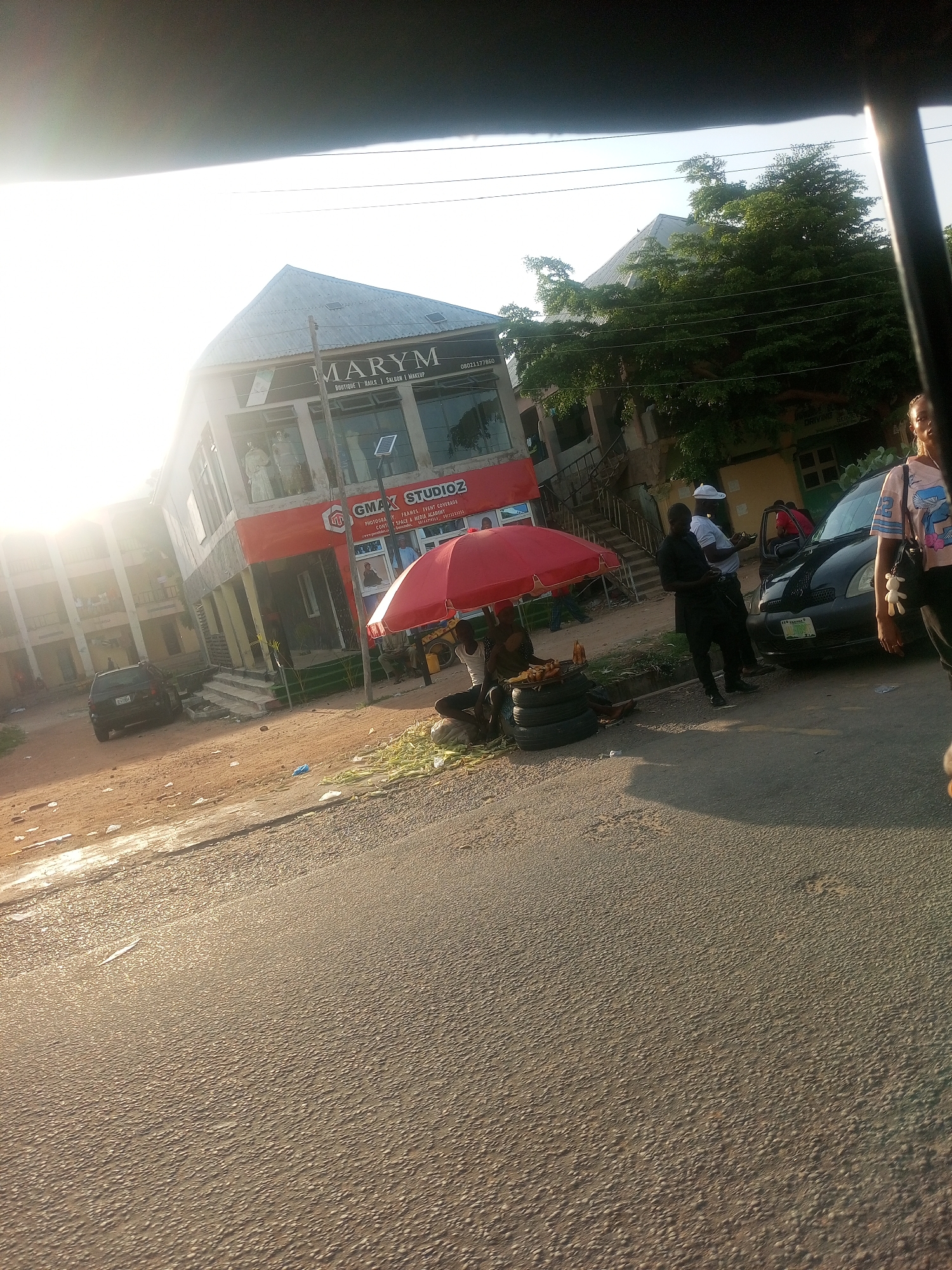 In Jalingo, Nigeria, a street scene unfolds under the warm glow of what appears to be late afternoon sun. The focal point is a two-story building with a modern design, housing businesses named "MARYM" and "GMAX STUDIOZ." "MARYM" advertises itself as a boutique, nail salon, and makeup service, with a visible phone number: 08021177860. Below it, "GMAX STUDIOZ" offers photography, frames, and event coverage, with mention of a "Space & Media Academy."
In front of the building, a red umbrella shades a small vendor's stall. Underneath, several stacked tires serve as a display base for items, possibly food or goods, with a bottle of liquid present. A few people are gathered around this area, with one individual seated and another standing.
Further down the street, a black car is parked. To the right of the car, a man in dark clothing stands while another, wearing a white hat, leans in towards the car. A woman in a light-colored t-shirt with a graphic print is visible on the far right edge of the frame, facing towards the building. Adjacent to the building on the left, another multi-story structure with balconies is visible, suggesting a busy urban environment. The ground appears to be a mix of dirt and asphalt, with some debris scattered around. The overall impression is of a bustling commercial area on a clear day, with signs of everyday commerce and social interaction.
